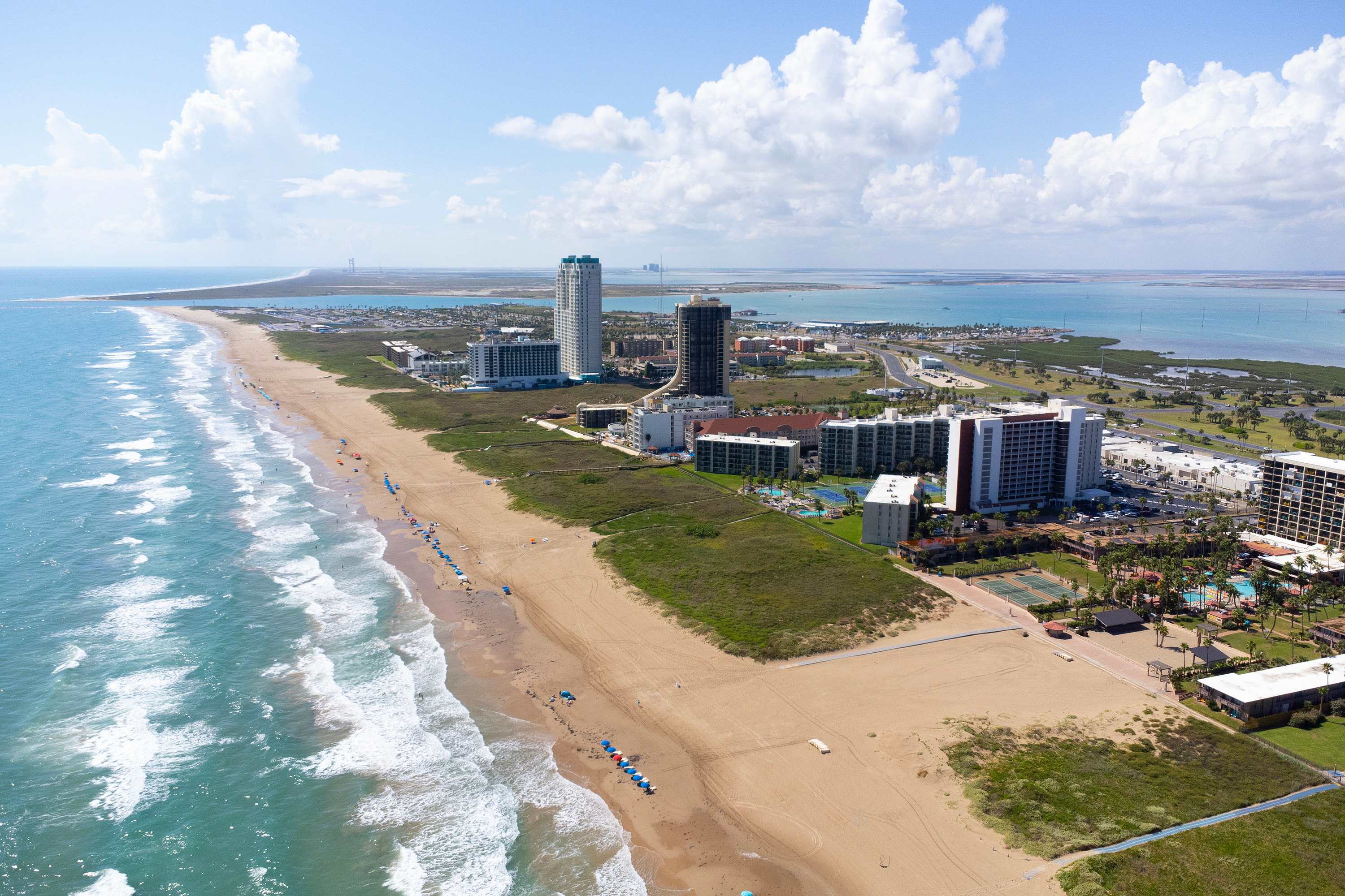 An expansive aerial view of the coastal area, showing the long sandy beach, the blue ocean, numerous high-rise buildings, and the bay with distant landmasses under a partly cloudy sky.