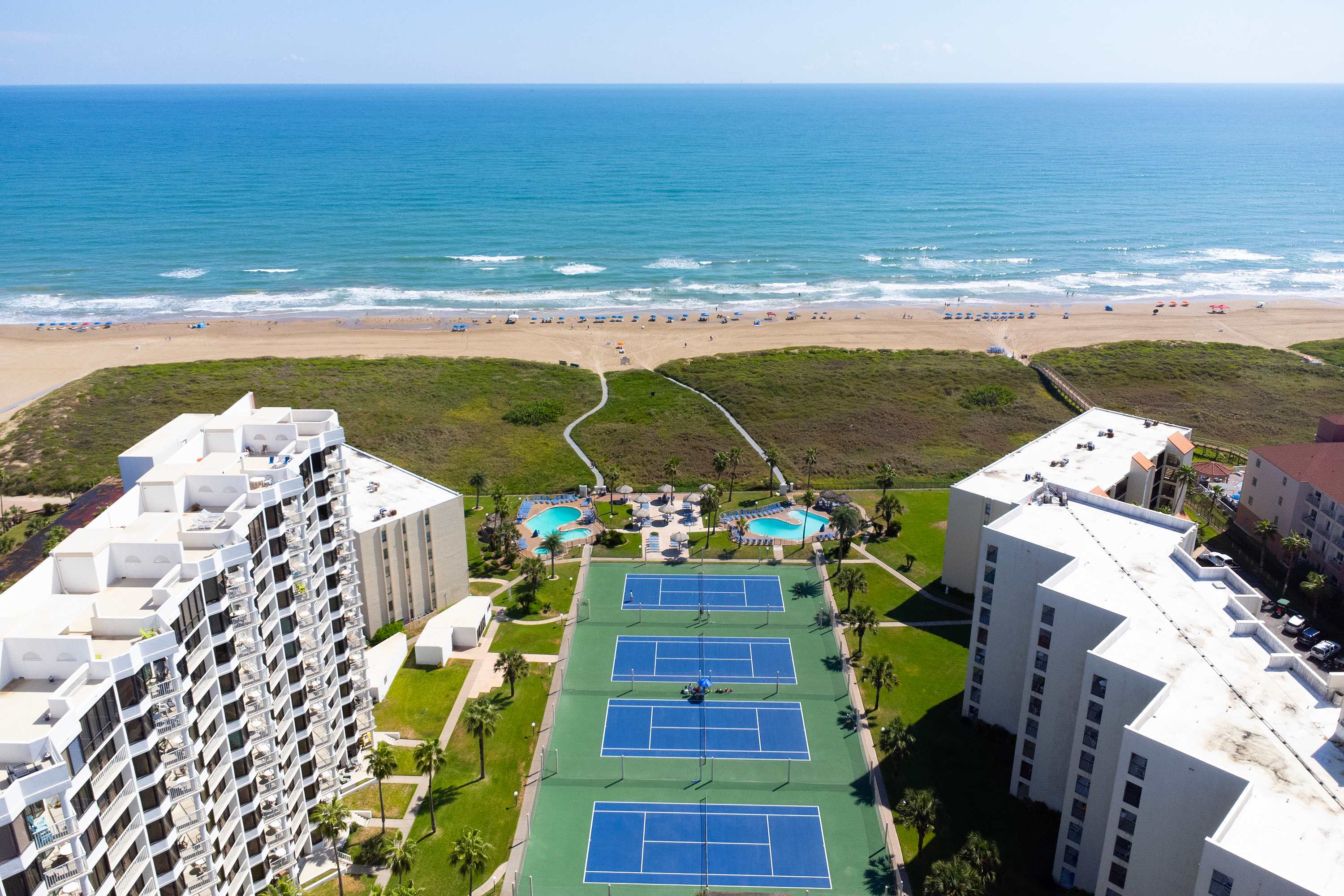 A wide aerial shot capturing the beachfront property with its buildings, tennis courts, and pools, set against the expansive blue ocean and sandy beach, extending along the coastline under a clear sky.