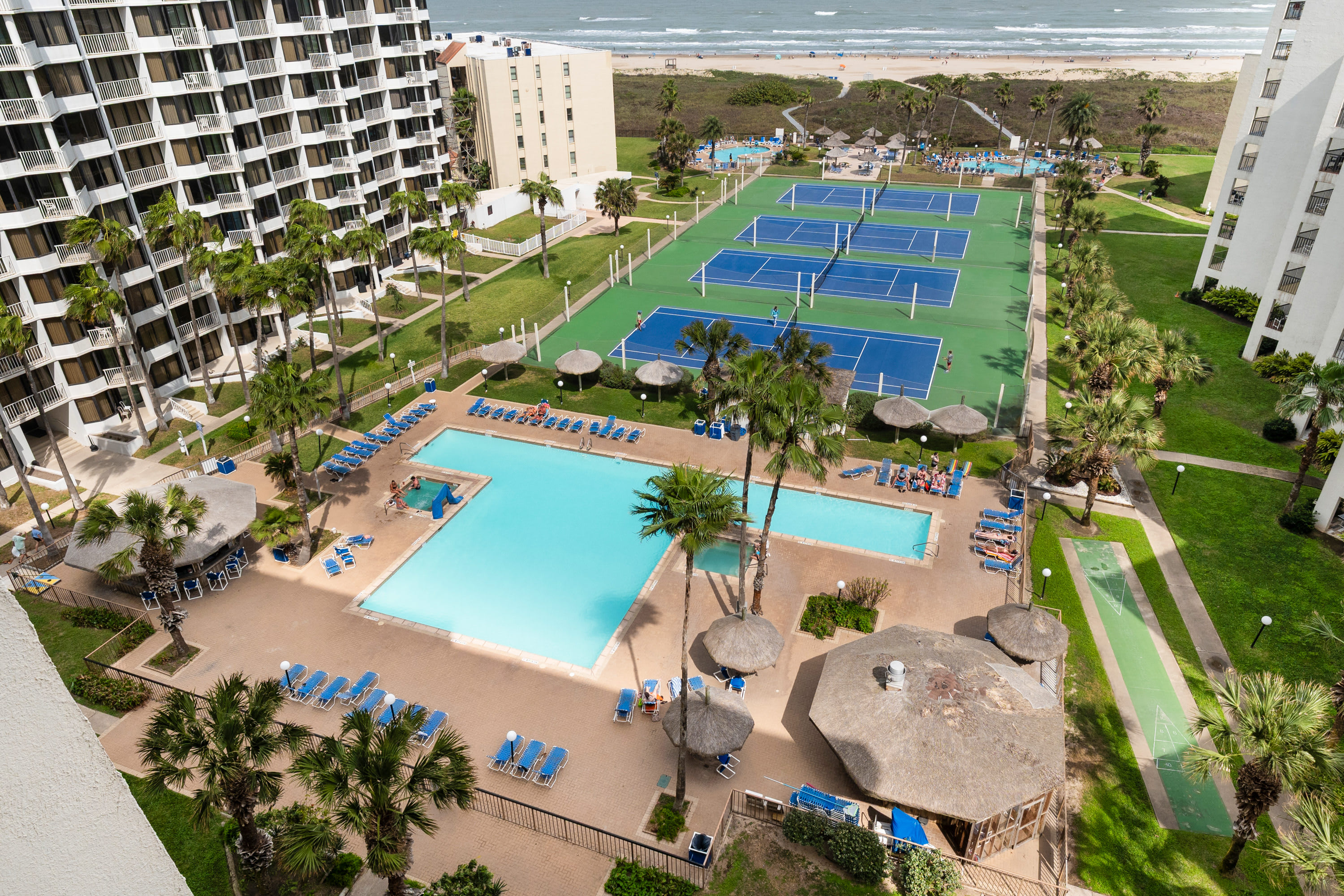 An aerial view showcasing the property's extensive outdoor amenities, including two large swimming pools, multiple tennis courts, lush green lawns with palm trees, and the ocean beach in the background.