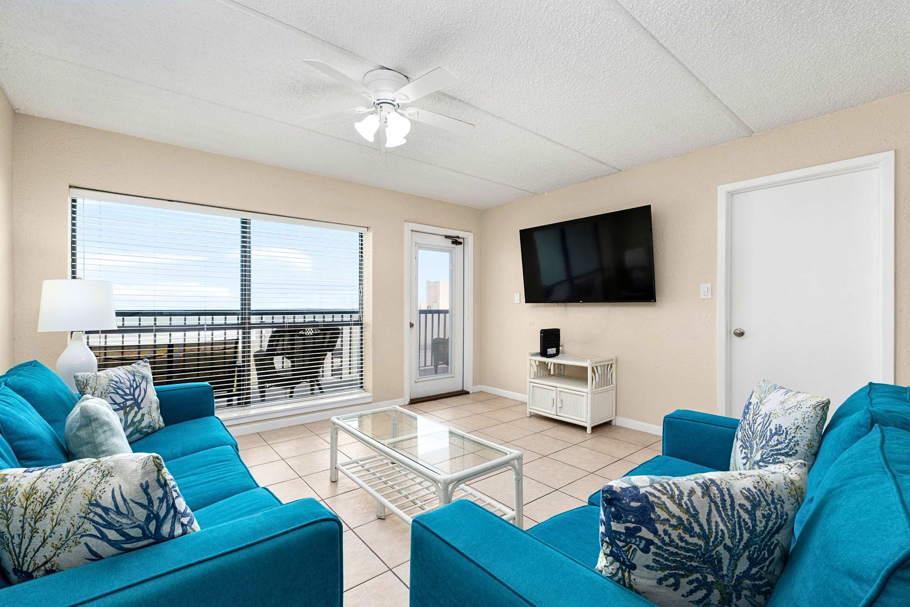 Inviting living room with two blue sofas, a glass coffee table, and a wall-mounted flat-screen TV above a white console, with a door leading to the balcony and ocean views.