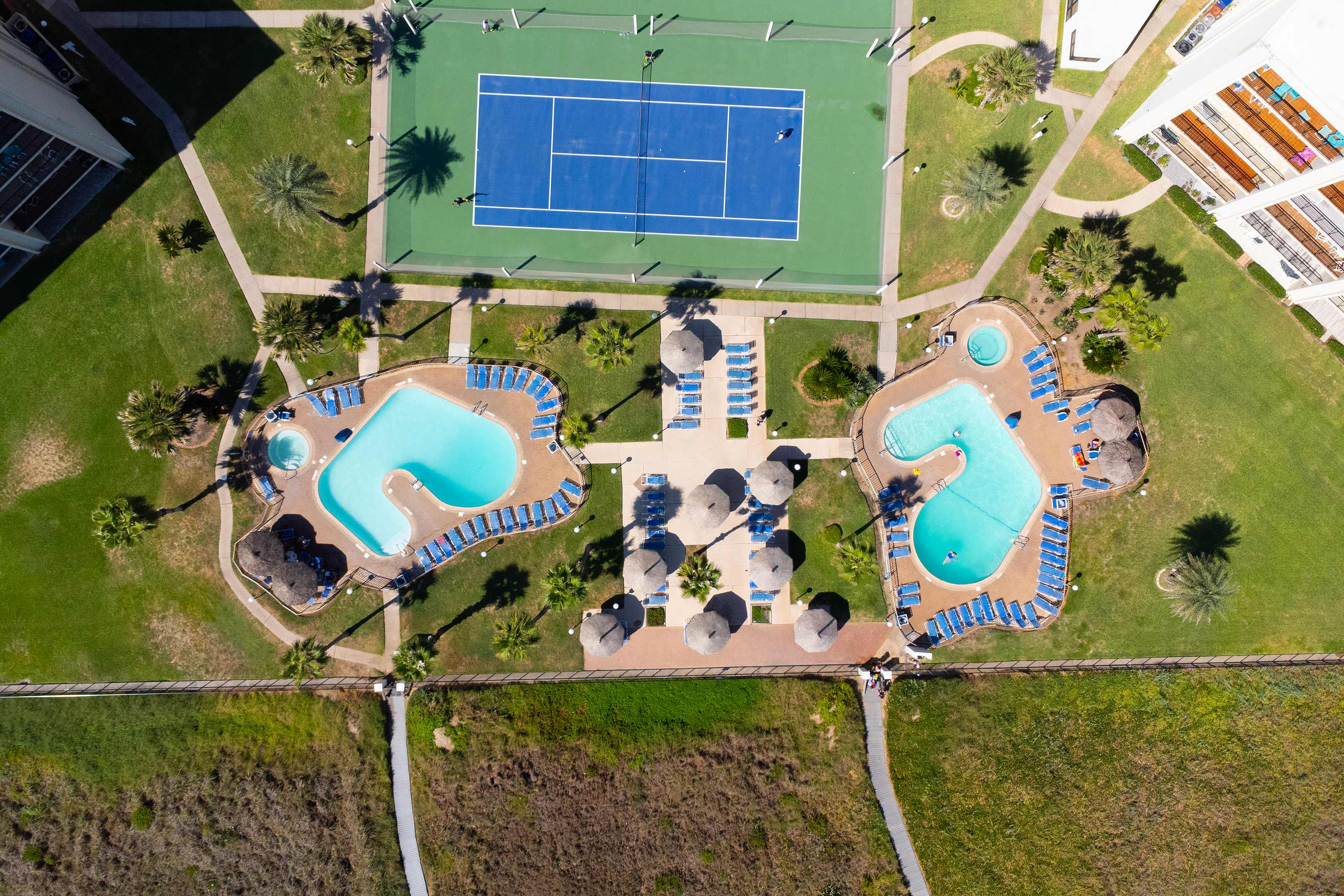 A top-down aerial view highlighting the property's recreational area with two distinct swimming pools surrounded by lounge chairs and umbrellas, adjacent to multiple blue tennis courts and green pathways.