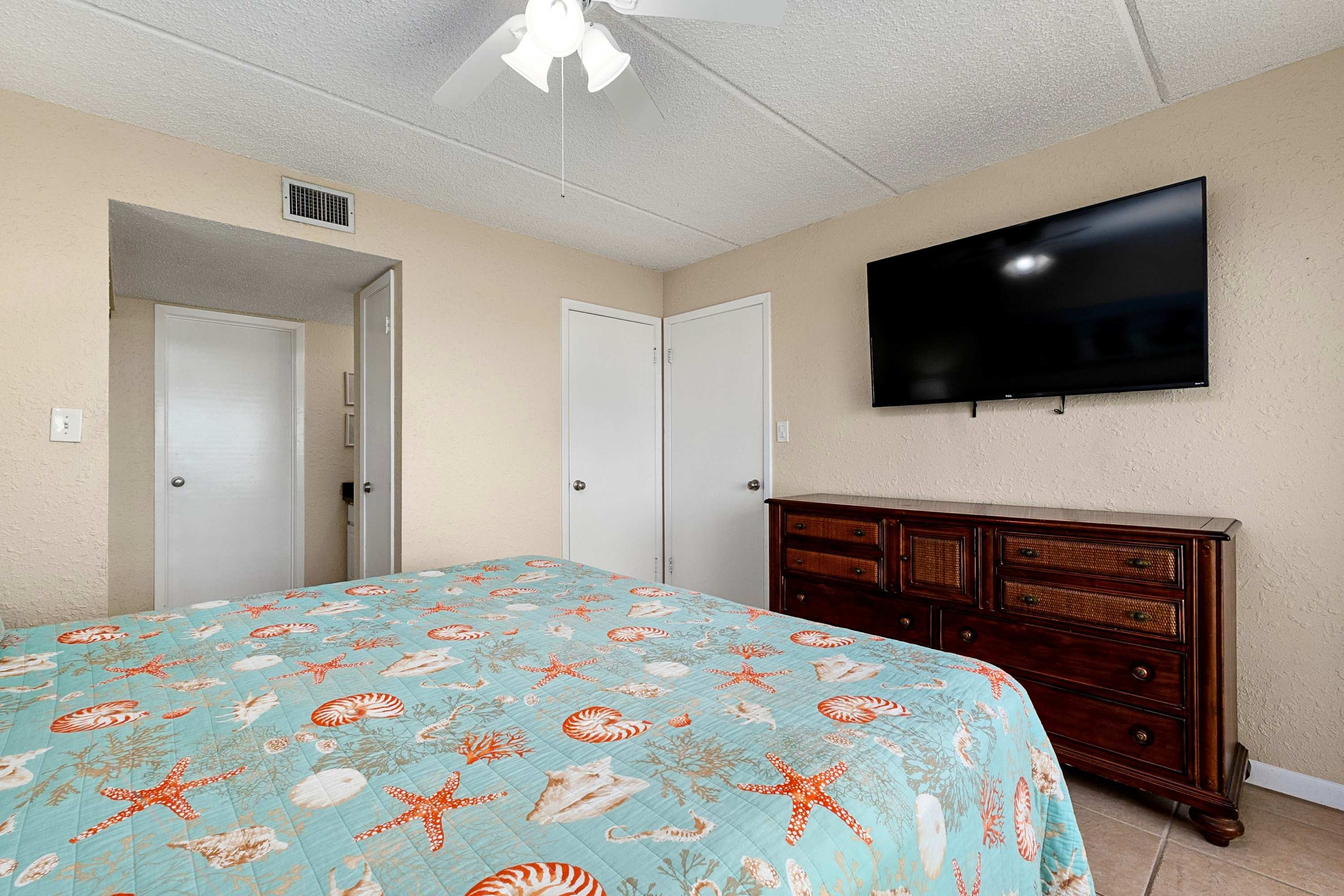 This bedroom features a bed with a vibrant blue and orange seashell-patterned comforter, a wall-mounted flat-screen TV, a dark wood dresser with wicker accents, and a white ceiling fan.
