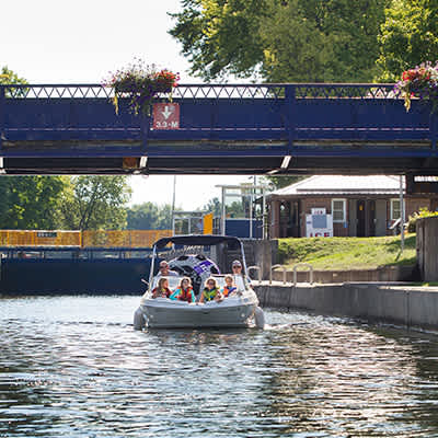 A pontoon boat filled with people passes under a blue metal bridge with hanging flower baskets, navigating a calm waterway with a building visible on the bank.