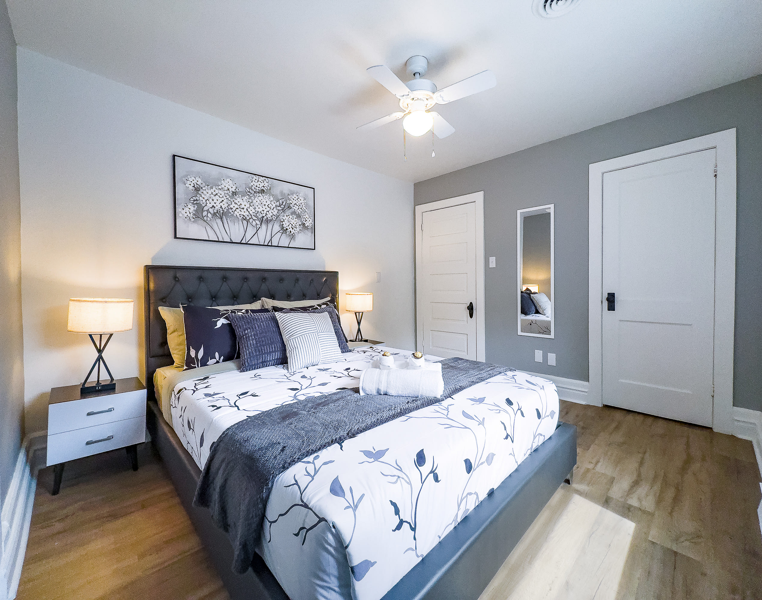 A cozy bedroom with light wood floors and gray accent walls, featuring a dark gray tufted headboard on a queen bed with white and gray floral bedding. Two nightstands with lamps flank the bed, and a white ceiling fan is overhead.