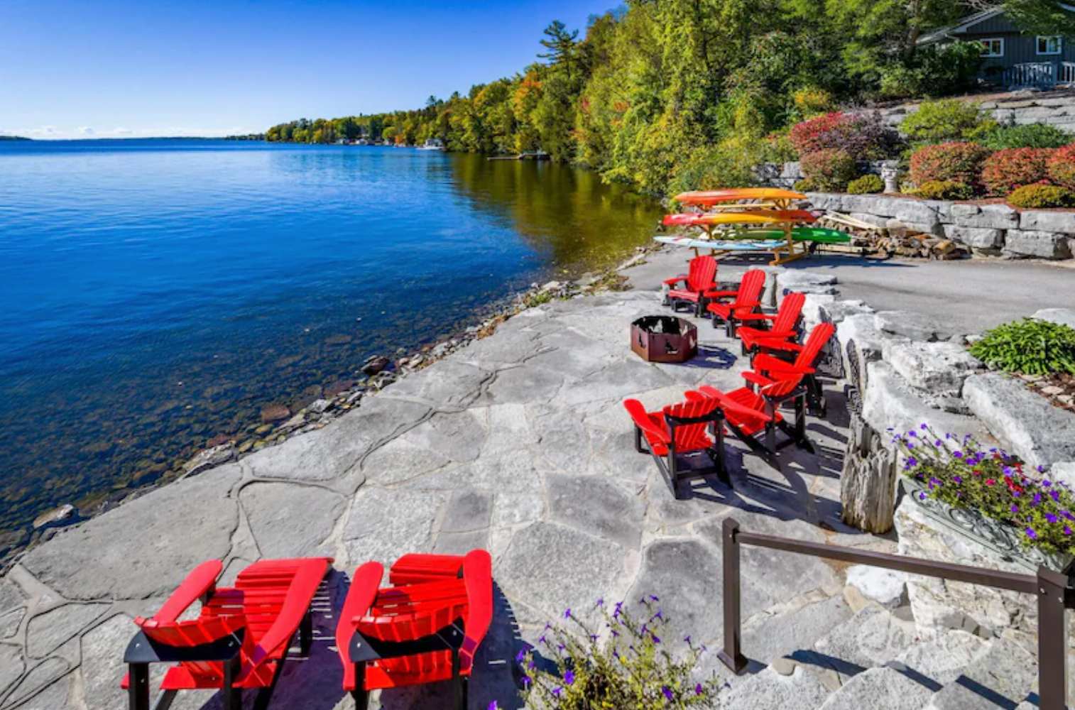 A scenic lakeside area features a stone patio with multiple red Adirondack chairs and a fire pit, overlooking clear blue water and a forested shoreline with kayaks.