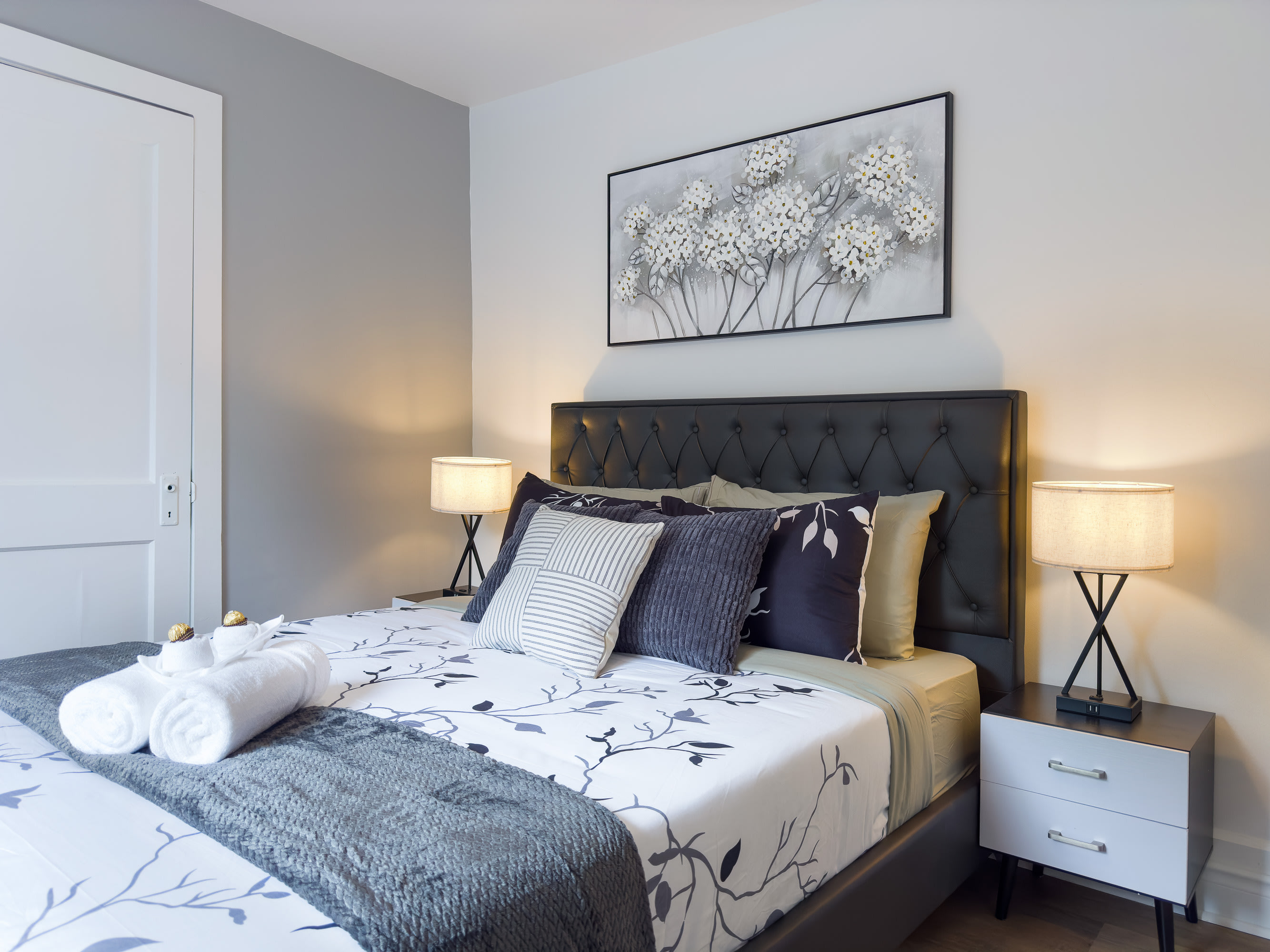 A close-up of the queen bed in bedroom 1, featuring a dark gray tufted headboard, white and gray floral bedding, and multiple decorative pillows. White rolled towels are neatly placed on the bed, flanked by nightstands with lamps.