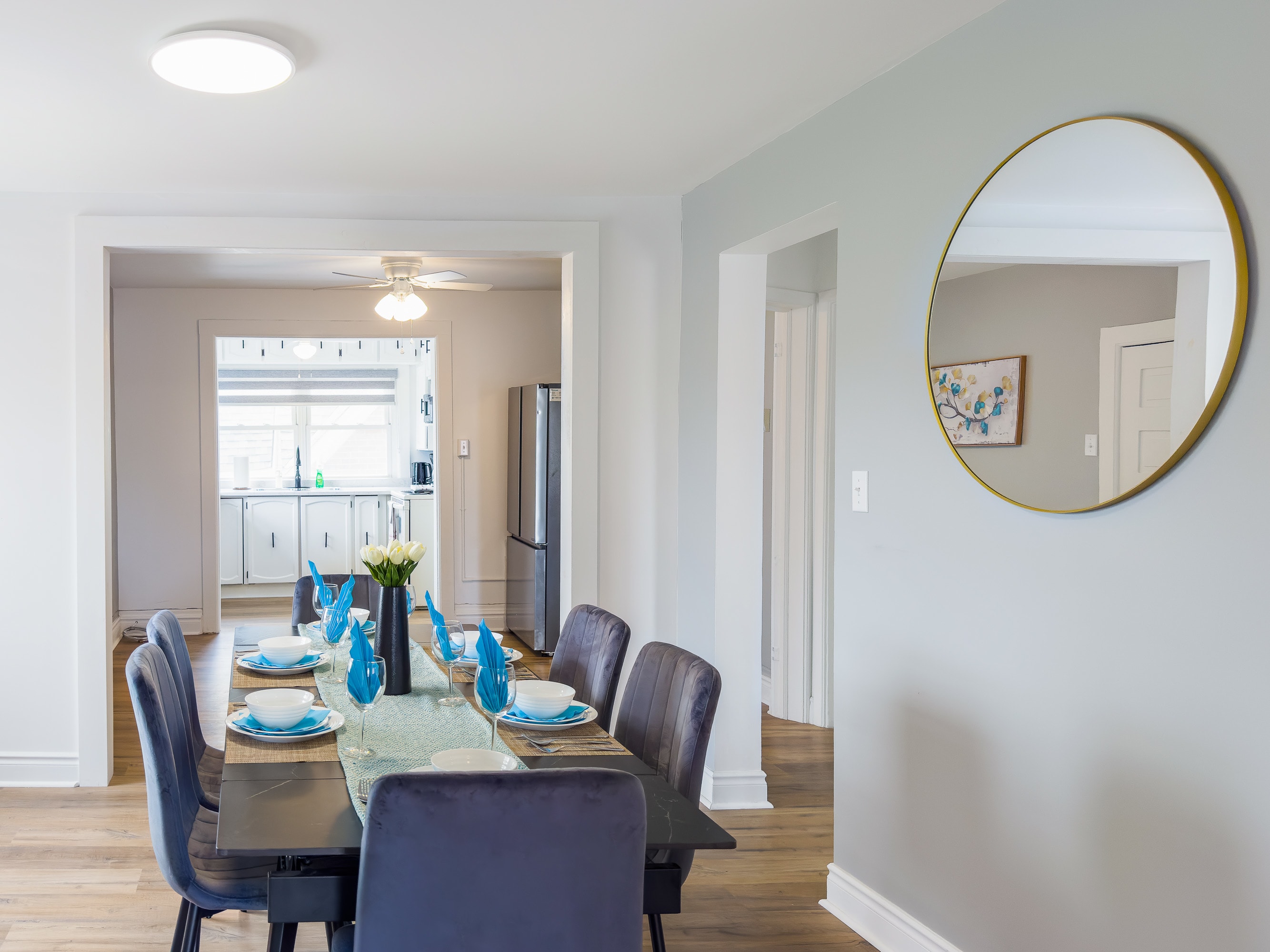 A dining area with light gray walls and wood flooring features a dark rectangular dining table set with blue and white dishes, surrounded by gray velvet chairs. The kitchen is visible through an archway in the background, and a round gold-framed mirror hangs on the wall.