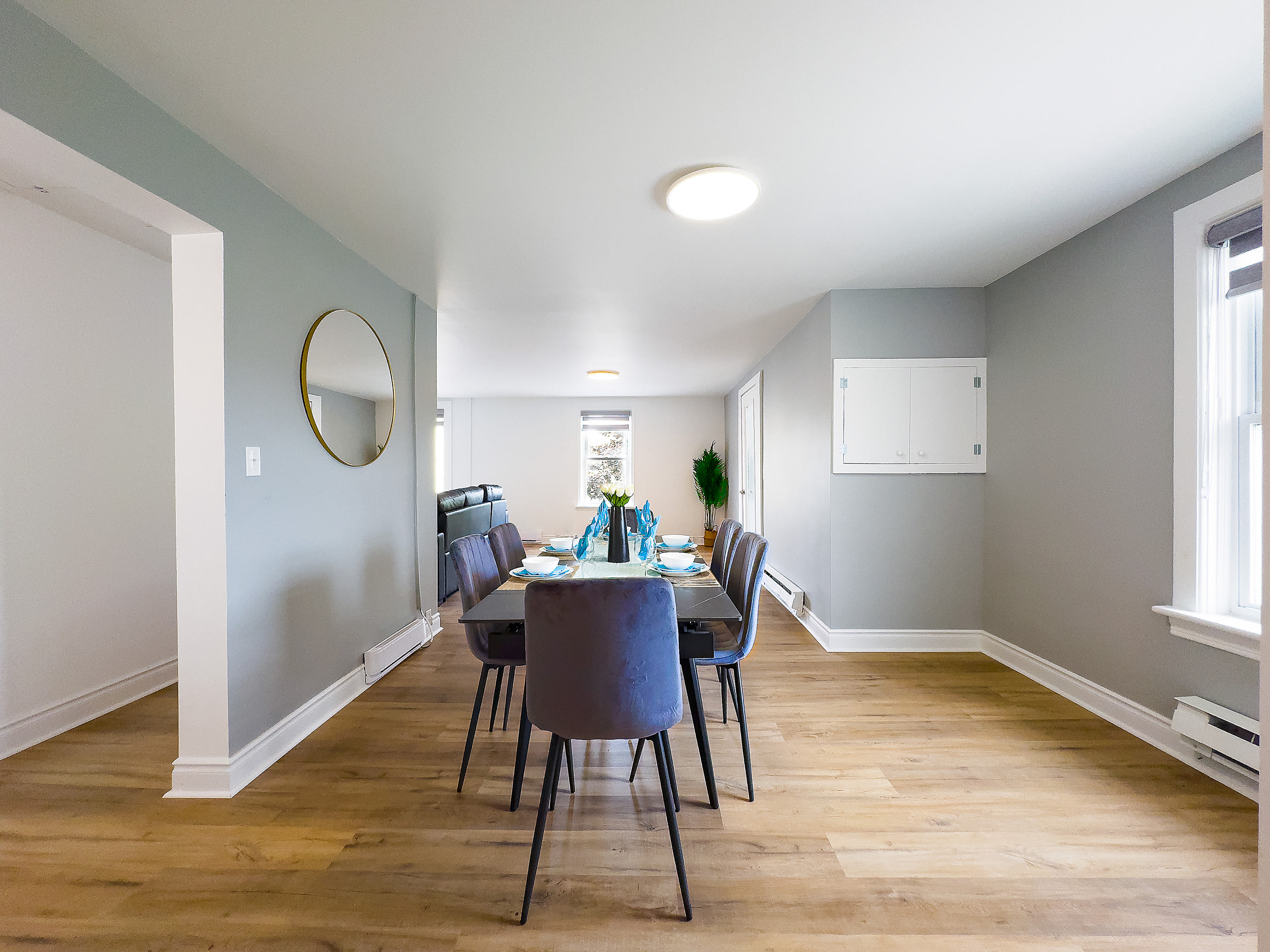 A spacious dining room features a dark rectangular dining table with six gray velvet chairs, set on light wood flooring. The light gray walls are adorned with a round gold-framed mirror, and a large window provides natural light.