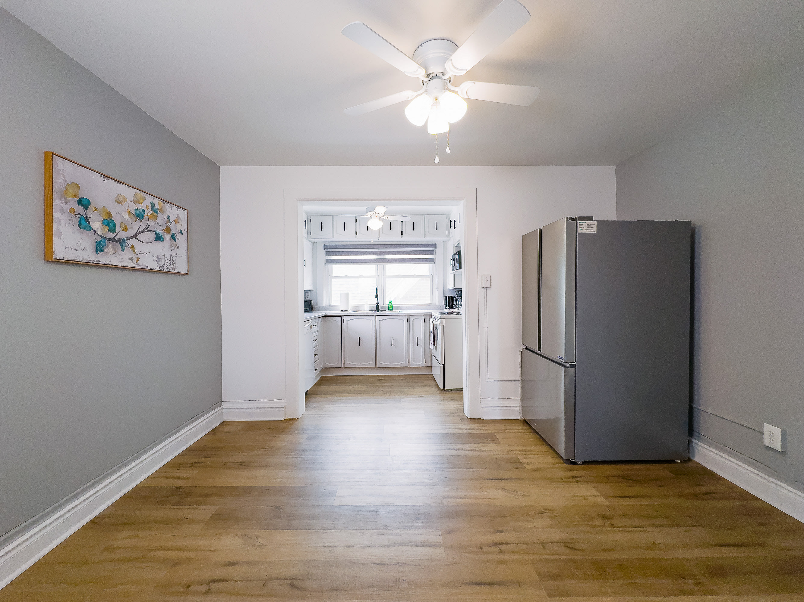 A bright room with light wood floors and gray walls, leading into a white kitchen with a window above the sink. A large stainless steel refrigerator stands to the right, and a white ceiling fan hangs overhead.
