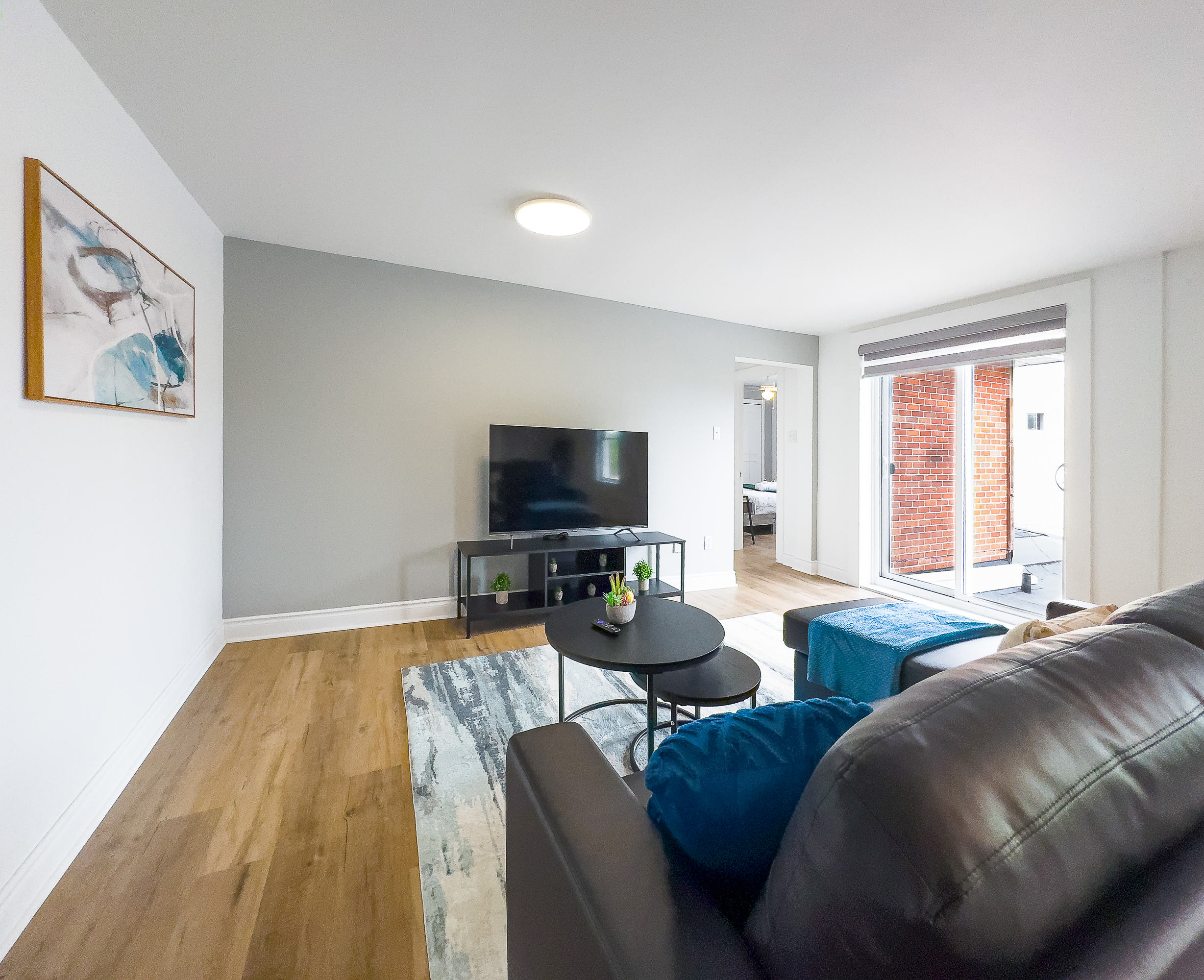 A modern living room features a black sectional sofa with blue accent pillows and a blue throw, facing a large flat-screen TV on a black console. A nested black coffee table sits on a blue and white abstract rug, with light wood flooring and a sliding glass door to the right.