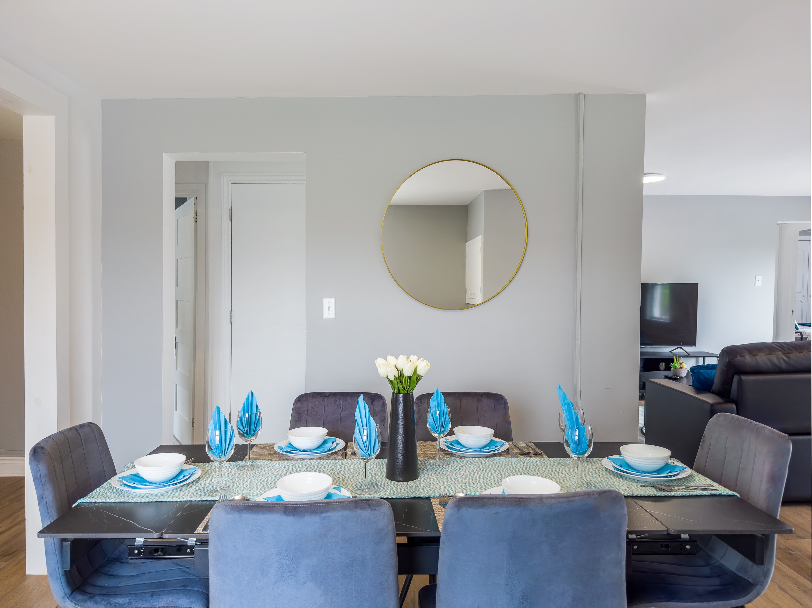 A modern dining room features a black rectangular table set for six with blue and white place settings, a teal runner, and a white tulip centerpiece. A round gold-framed mirror hangs on the light gray wall, reflecting a living area.