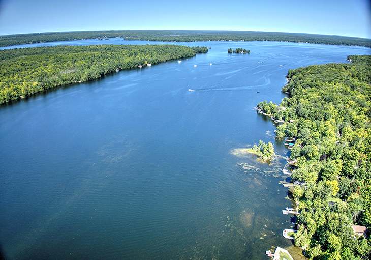 An aerial view captures a vast blue lake surrounded by dense green forests under a clear sky, with several small islands and boats visible on the water.