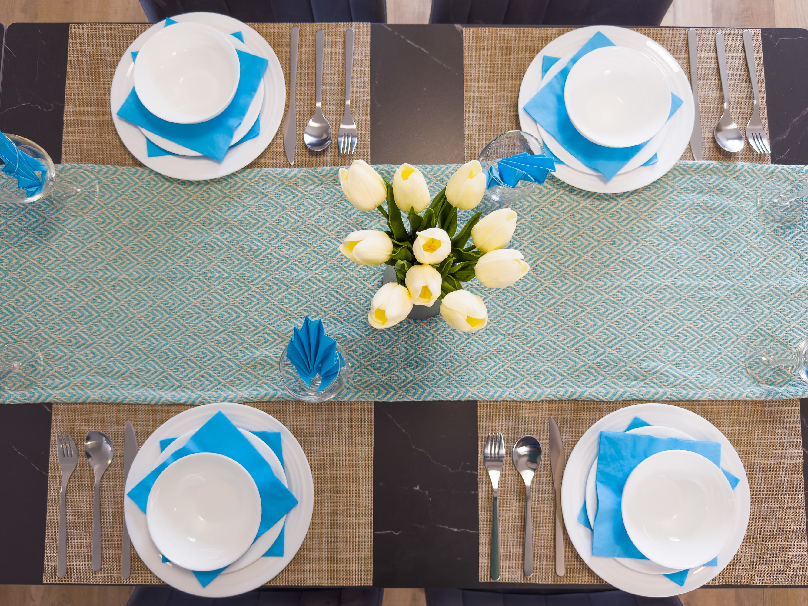 An overhead view of a modern dining table set for six, featuring a teal patterned runner, white plates, blue napkins, and a white tulip centerpiece. Each place setting includes a white bowl, plate, and silverware on a woven placemat.