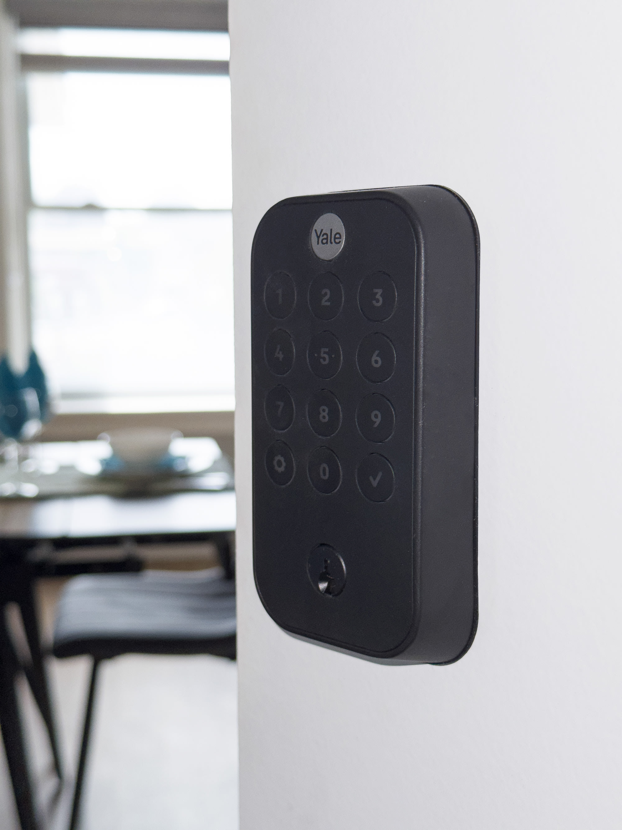 A black Yale smart lock with a keypad is mounted on a white wall, with a blurred view of a dining area and window in the background.