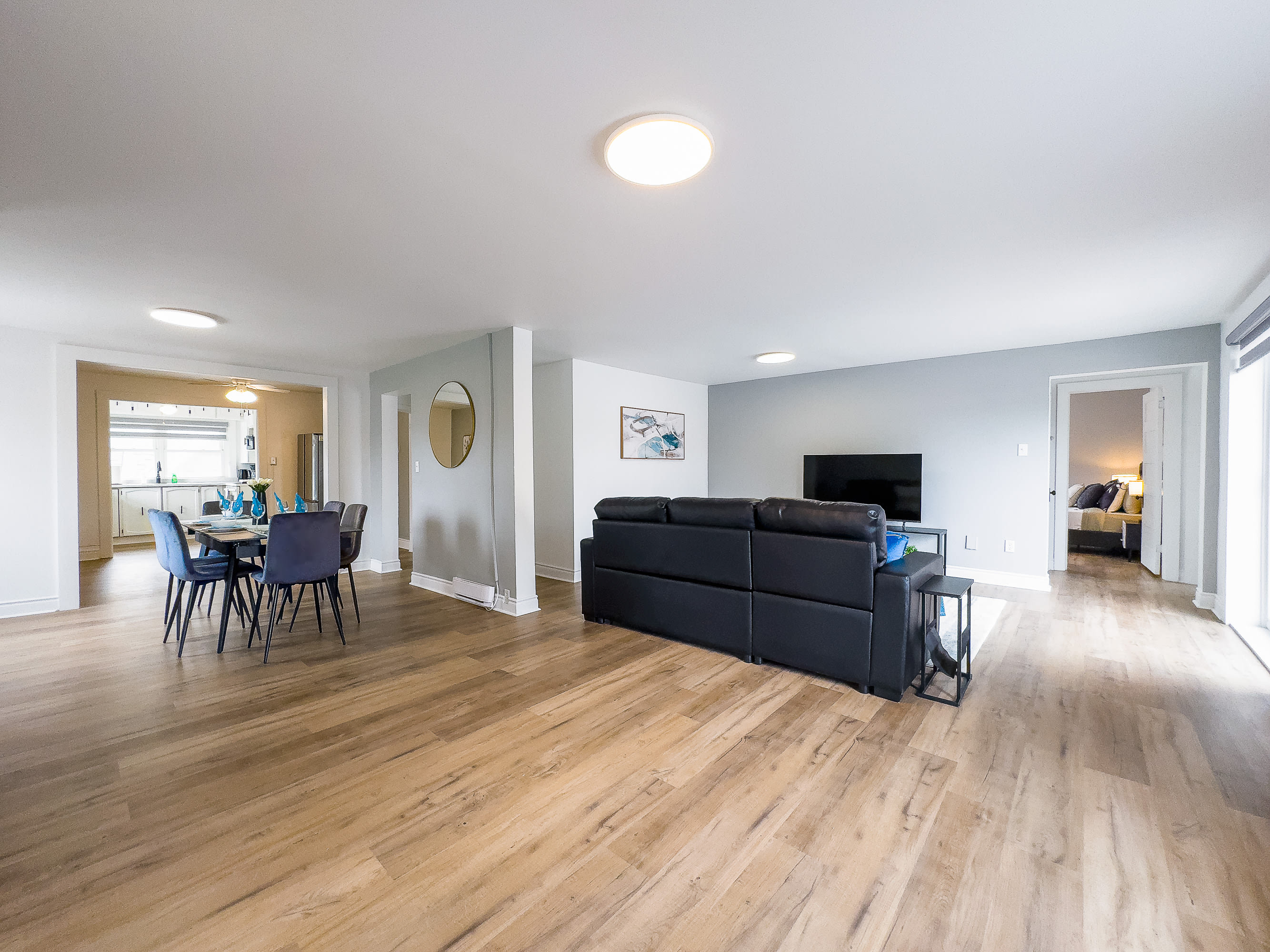 An open-concept living space features a black sectional sofa in the foreground, with a dining area visible to the left, furnished with a dark table and gray chairs. Light wood flooring extends throughout, connecting the living room to the dining area and a glimpse of the kitchen beyond.
