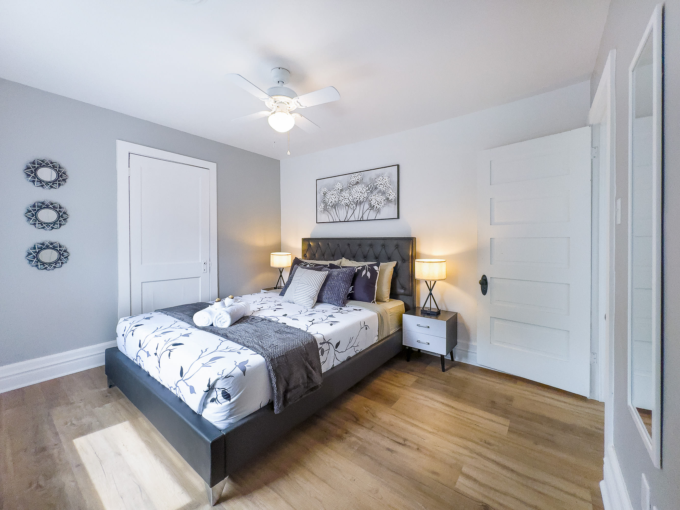 A wider view of the cozy bedroom with light wood floors and gray accent walls, showing a queen bed with dark gray tufted headboard and white floral bedding. Two white nightstands with lamps are on either side, and white doors are visible.