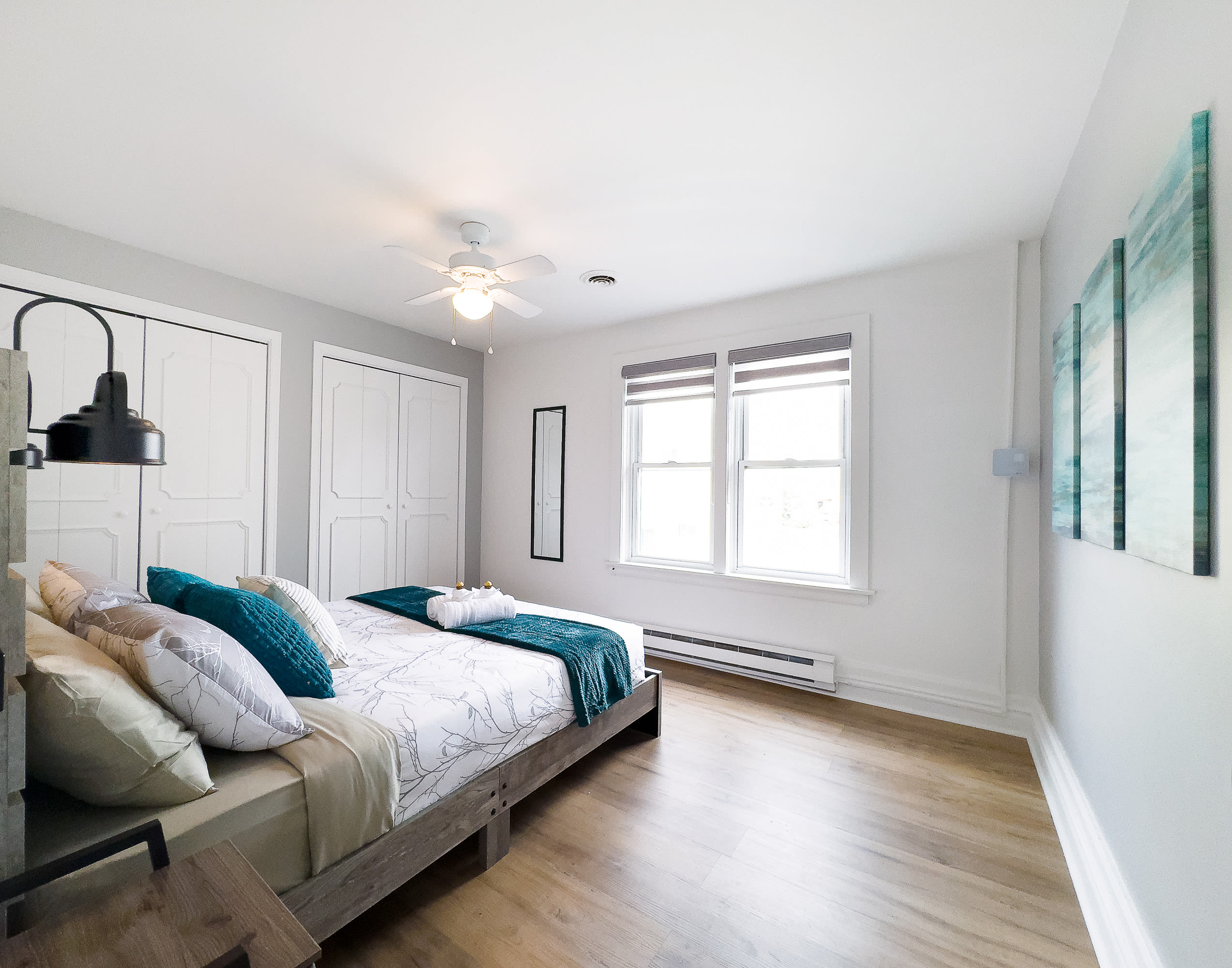 A wider view of bedroom 2, showing light wood floors, a queen bed with a rustic wooden headboard, and white and teal bedding. Two large windows with blinds provide natural light, and white closet doors are visible.