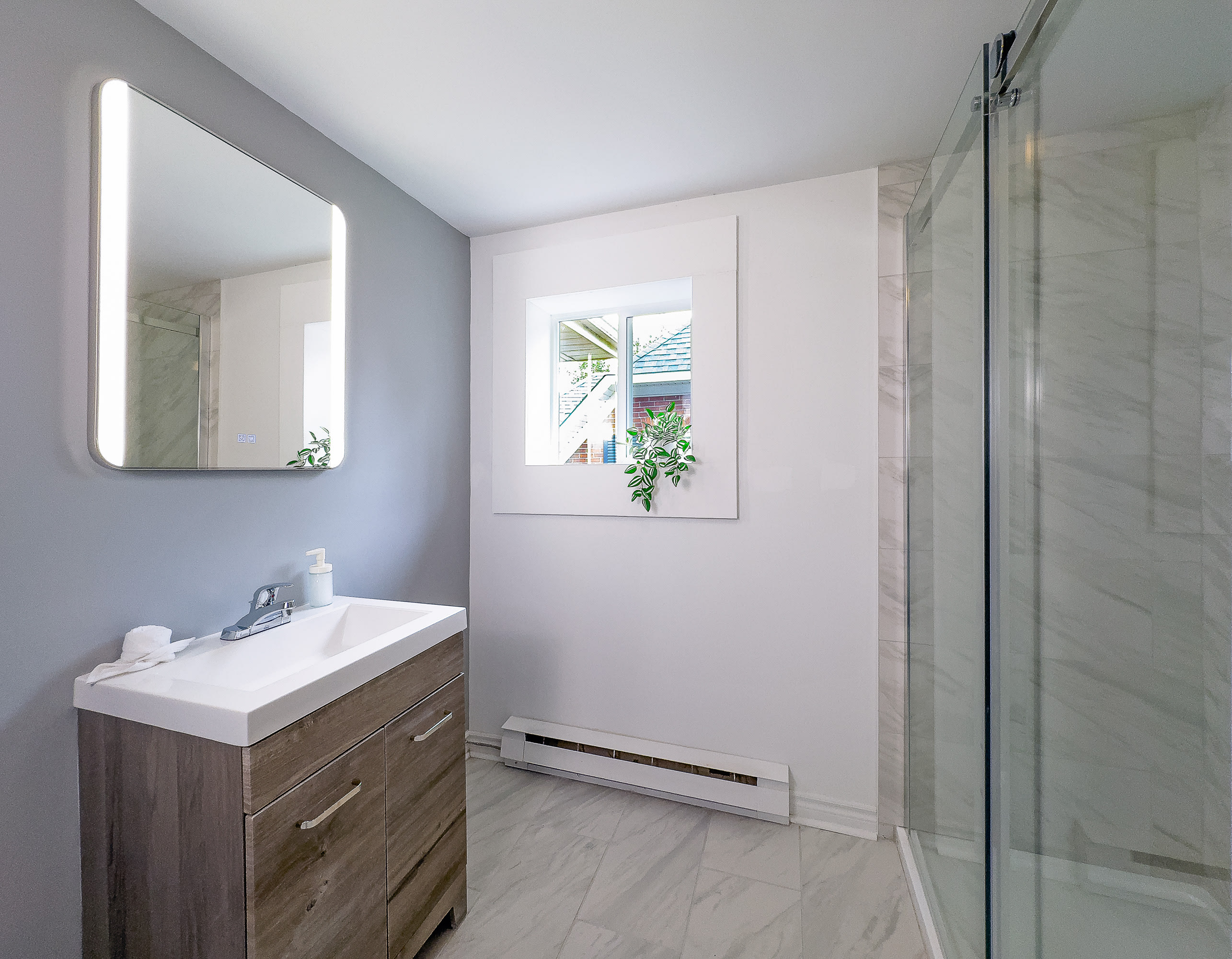 A modern bathroom features a light wood vanity with a white sink, a large illuminated mirror, and a glass-enclosed shower. The walls are painted light gray and white, complemented by light gray tiled floors.