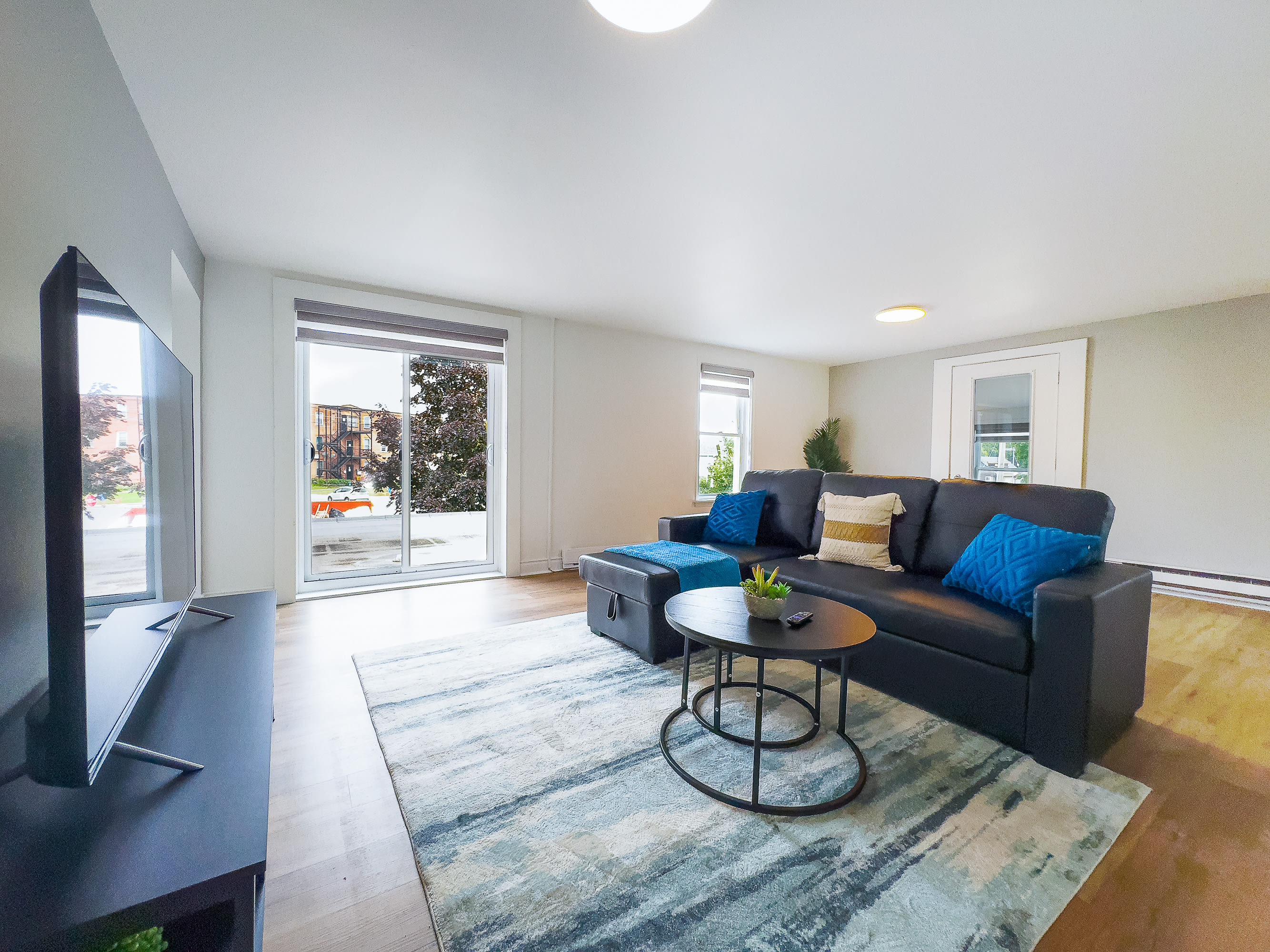 A spacious living room features a black sectional sofa with blue and beige accent pillows, and a blue throw blanket, facing a large flat-screen TV on a black console. A round black coffee table sits on a blue and white abstract rug, with light wood flooring and a sliding glass door leading to an outdoor area.