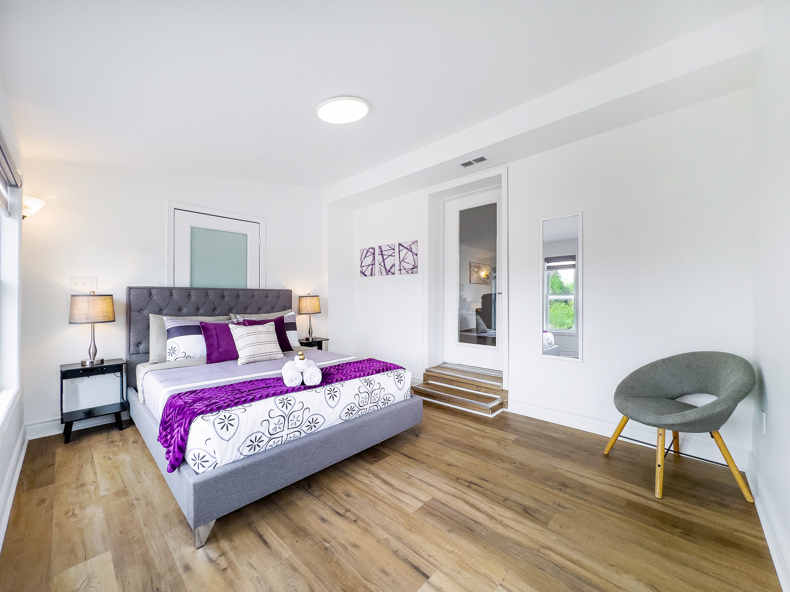 A bright bedroom features a gray upholstered bed with purple and white bedding, flanked by black nightstands with lamps. Hardwood floors extend across the room, leading to a raised doorway with a glass-paneled door.