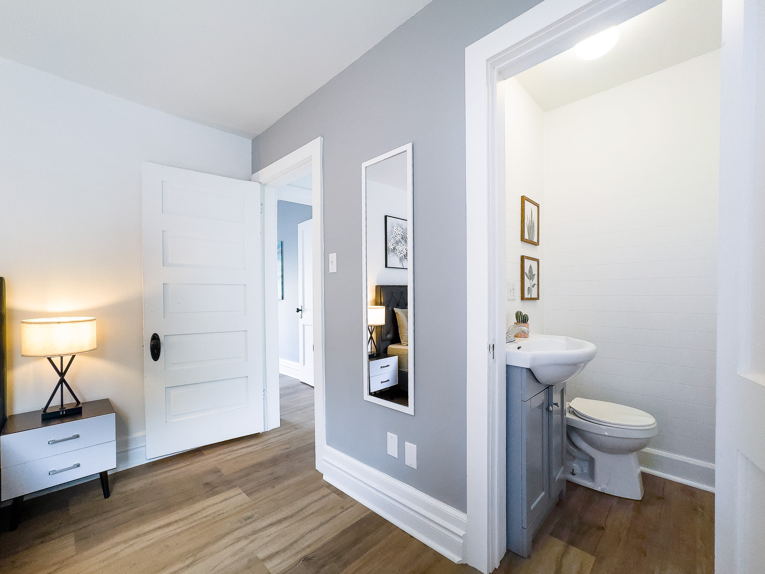 A view from bedroom 1 into a small powder room with a white sink, toilet, and light gray vanity. A full-length mirror hangs on the wall outside the bathroom, reflecting the bedroom.