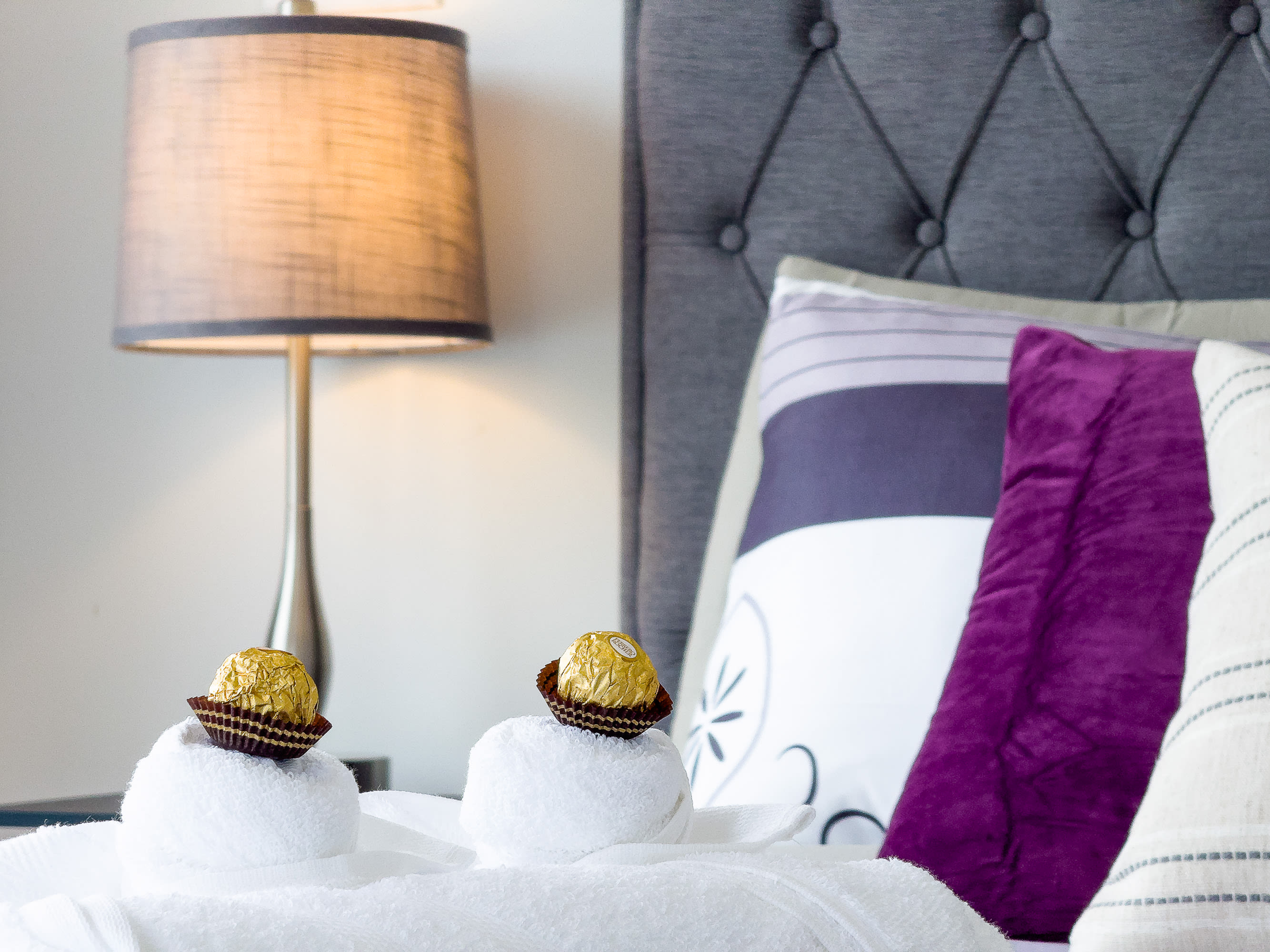 A close-up of the bed shows white rolled towels topped with chocolates, resting on purple and white patterned bedding. A gray tufted headboard and a bedside lamp are visible in the background.