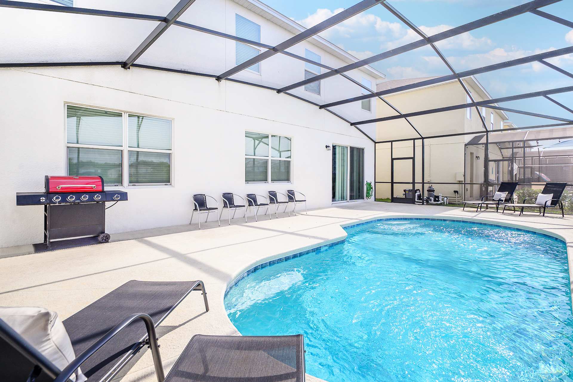 Screened-in private pool area showing the back of the white house with multiple windows and sliding glass doors. A red BBQ grill and additional seating are on the concrete deck next to the pool.