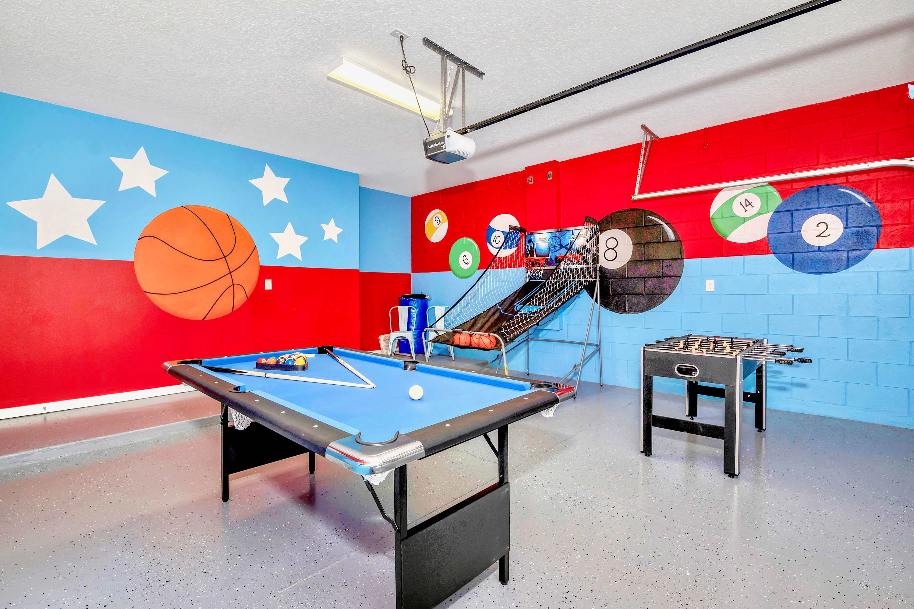 Spacious game room with vibrant red and blue walls adorned with large sports-themed murals, including a basketball and pool balls. It features a blue felt pool table, a foosball table, and a basketball arcade game.