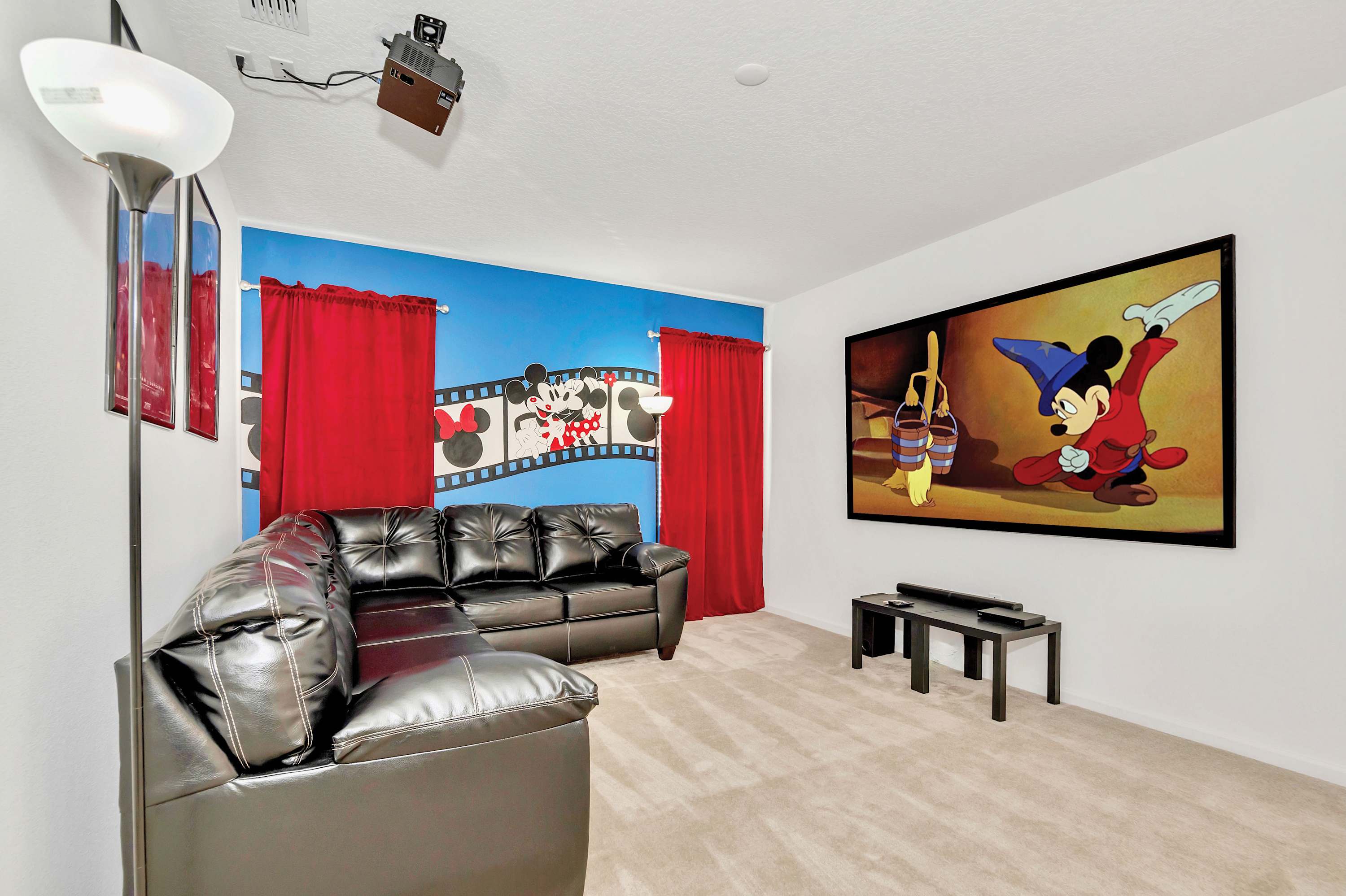 Cozy media room with a large projection screen displaying a Disney character, a black leather sectional sofa, and a blue accent wall with Mickey Mouse-themed decor and red curtains. The room has carpeted flooring and a floor lamp.