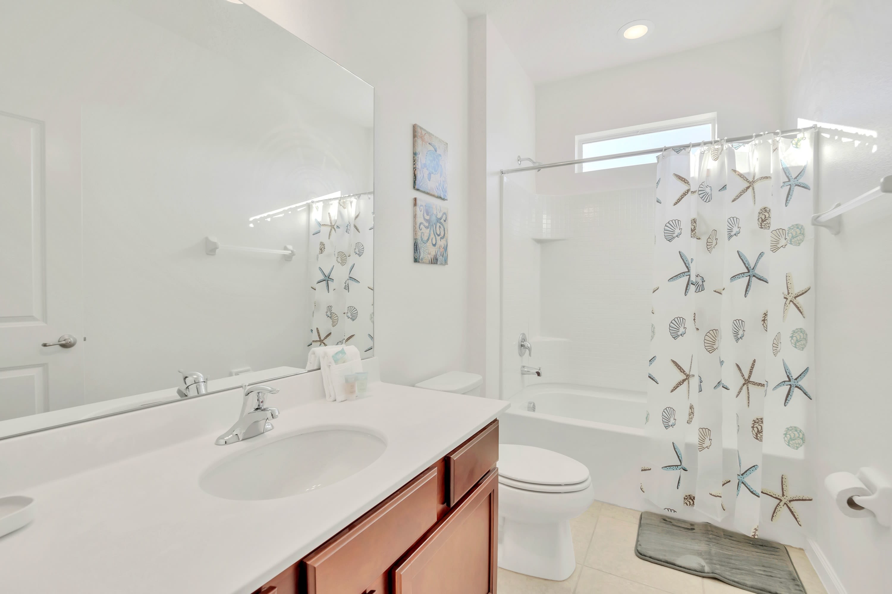 Well-lit bathroom featuring a white vanity with a single sink and wooden cabinets, a large mirror, and a toilet. A shower-tub combo with a nautical-themed curtain is visible, along with a small window above the tub.