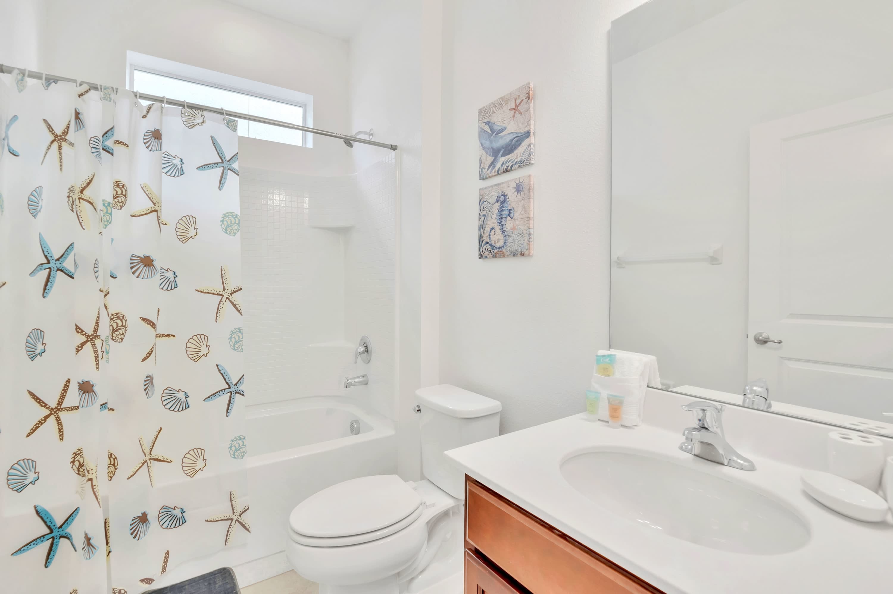Bright white bathroom featuring a shower-tub combo with a starfish and seashell-patterned curtain, a white toilet, and a vanity with a white countertop and wooden cabinets. Two framed nautical art pieces adorn the wall above the toilet.