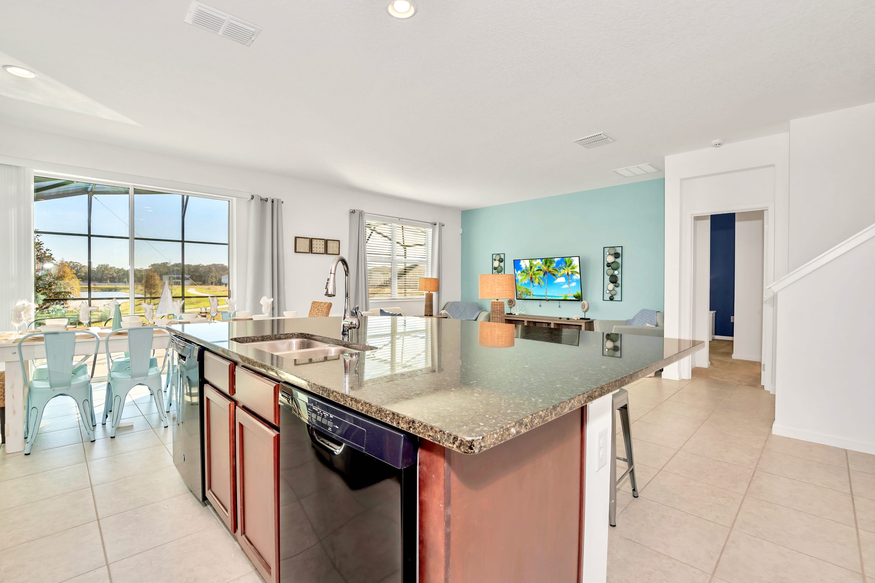 Open-concept view from the kitchen island, showcasing the seamless flow into the living room with a white sofa and TV, and the dining area with a view of the outdoor pool through large windows.
