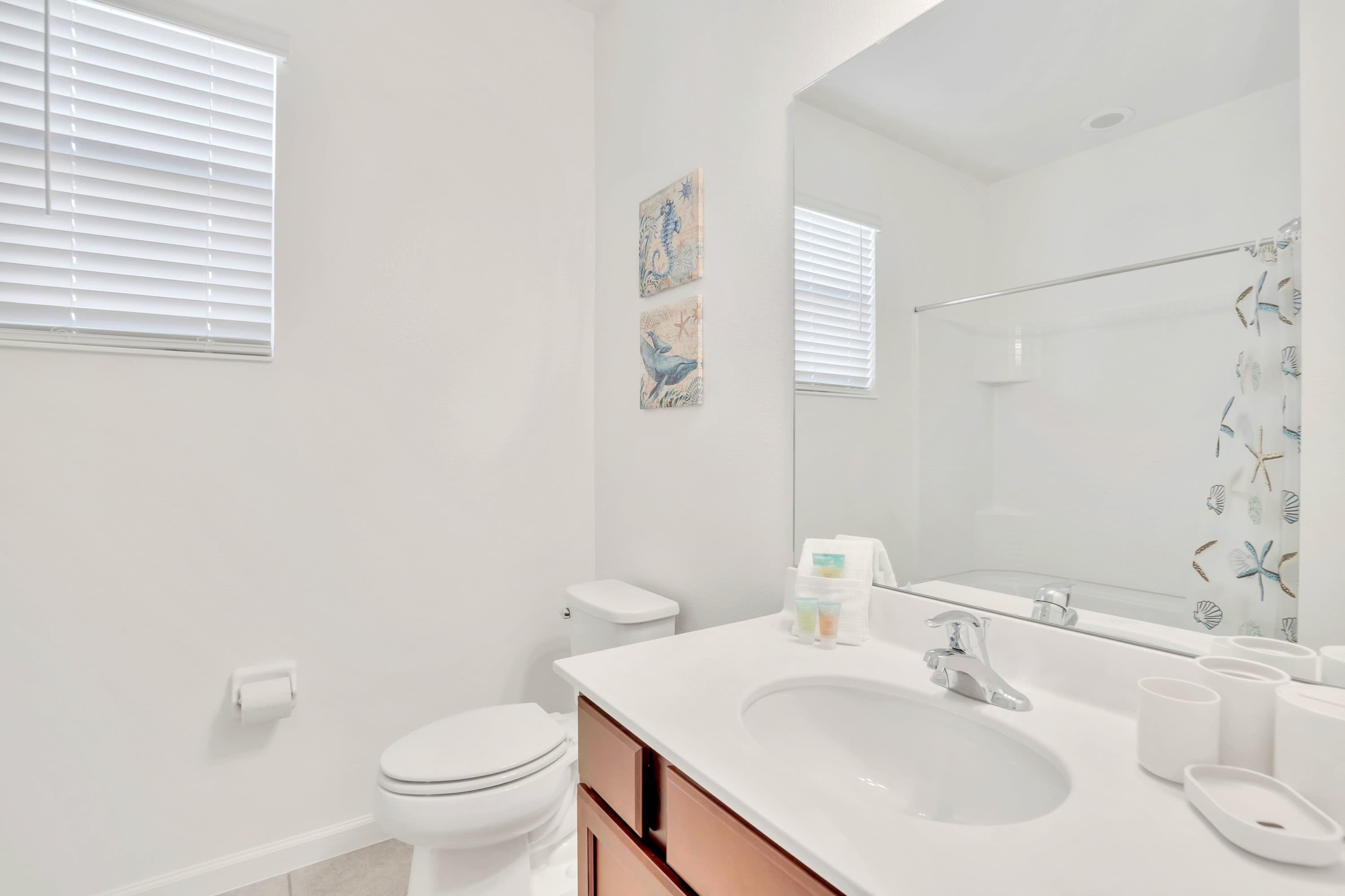 Clean white bathroom with a vanity featuring a white countertop and wooden cabinets, a large mirror, and a toilet. A window with blinds is visible, and two framed nautical artworks are on the wall.