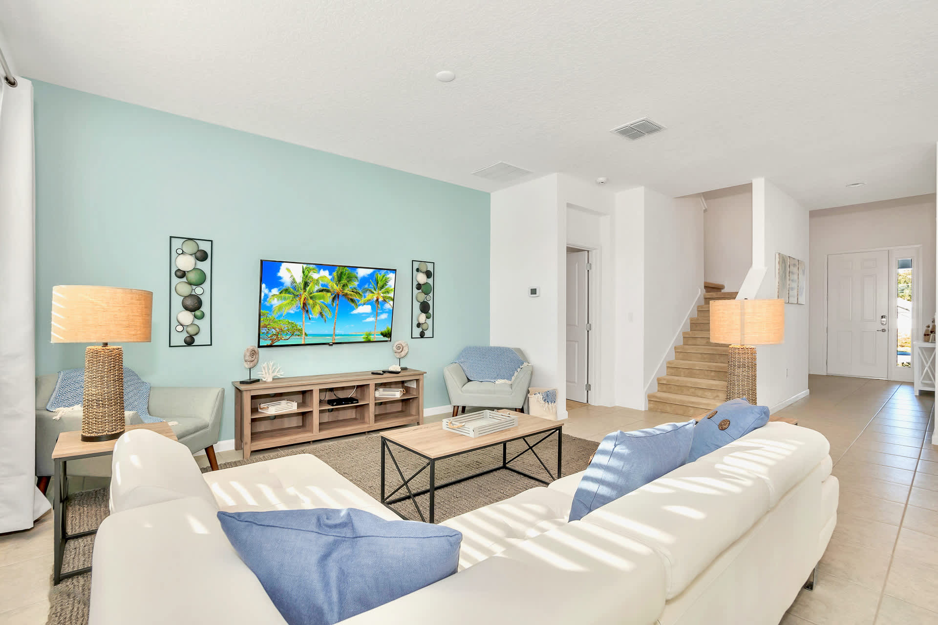 Bright living room with a light blue accent wall featuring a large TV, a white sectional sofa with blue pillows, a light wood coffee table, and two armchairs, all set on a neutral rug.