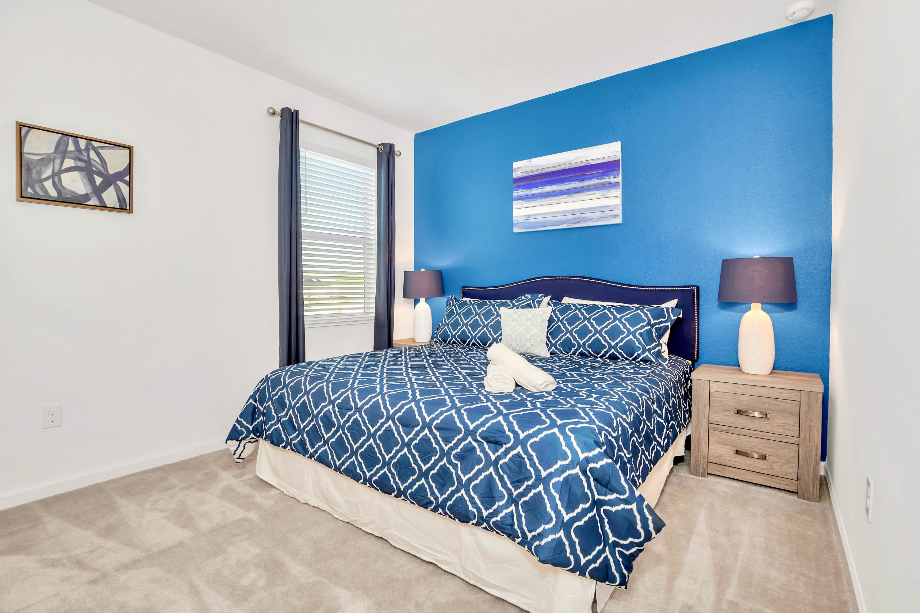 Modern bedroom with a striking blue accent wall behind a large bed featuring blue and white patterned bedding, flanked by two nightstands with lamps. A framed abstract artwork hangs above the headboard, and a window with blinds is visible.