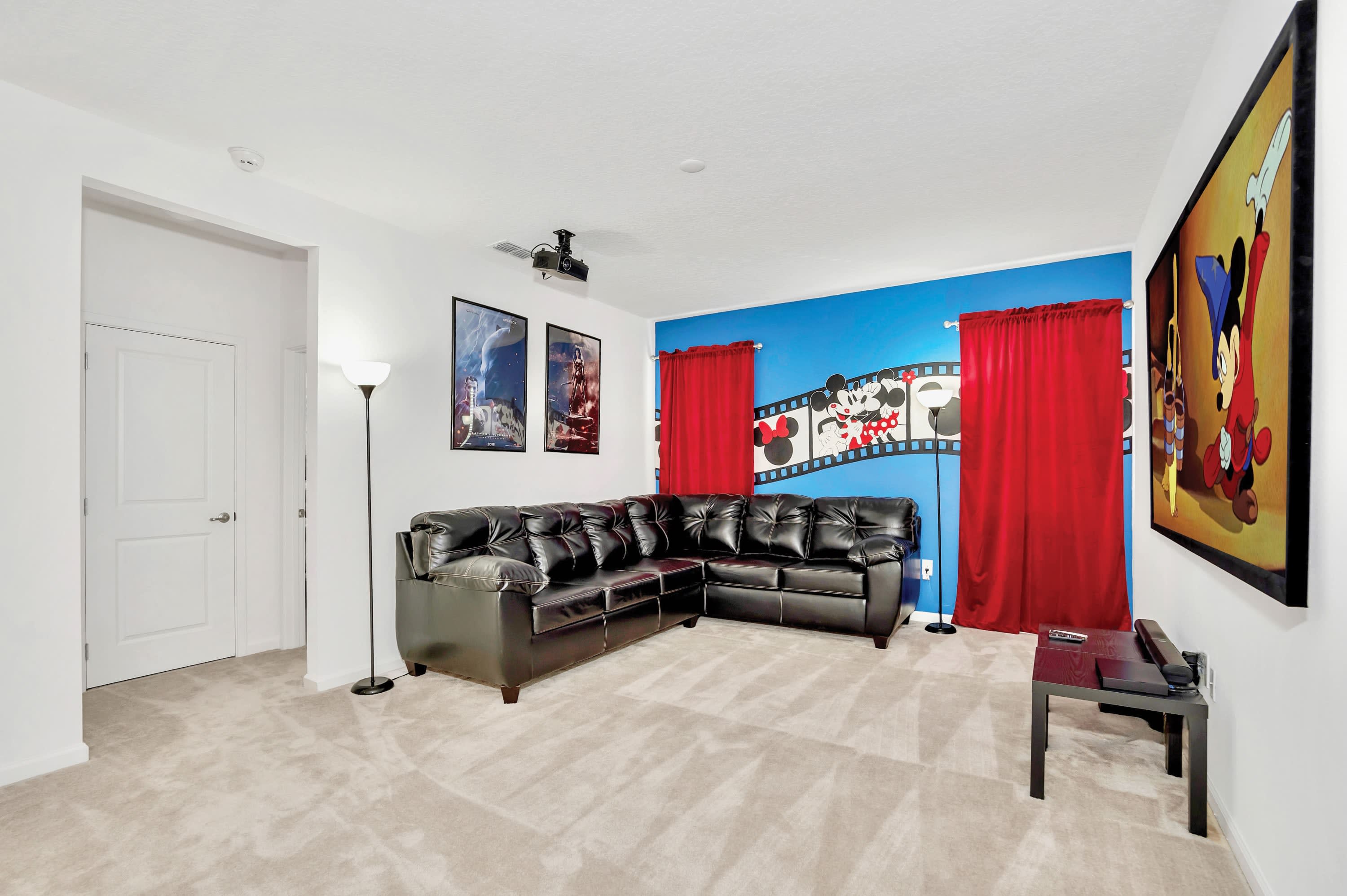 View of the media room featuring a large black leather sectional sofa facing a projection screen (partially visible). The room has a blue accent wall with Mickey Mouse decor, red curtains, and light carpeted flooring.