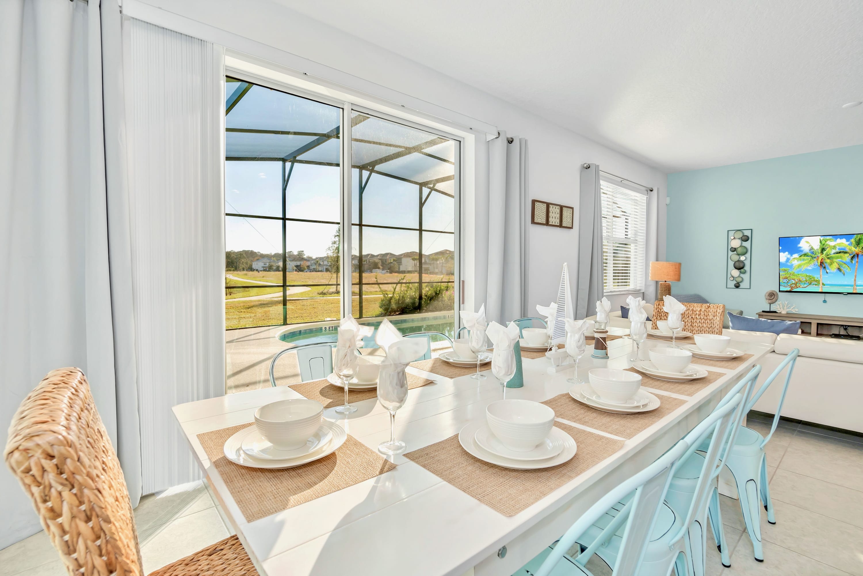 Elegant dining room featuring a long white table set for a meal, surrounded by light blue metal chairs and wicker end chairs, with large sliding glass doors opening to a screened pool area.