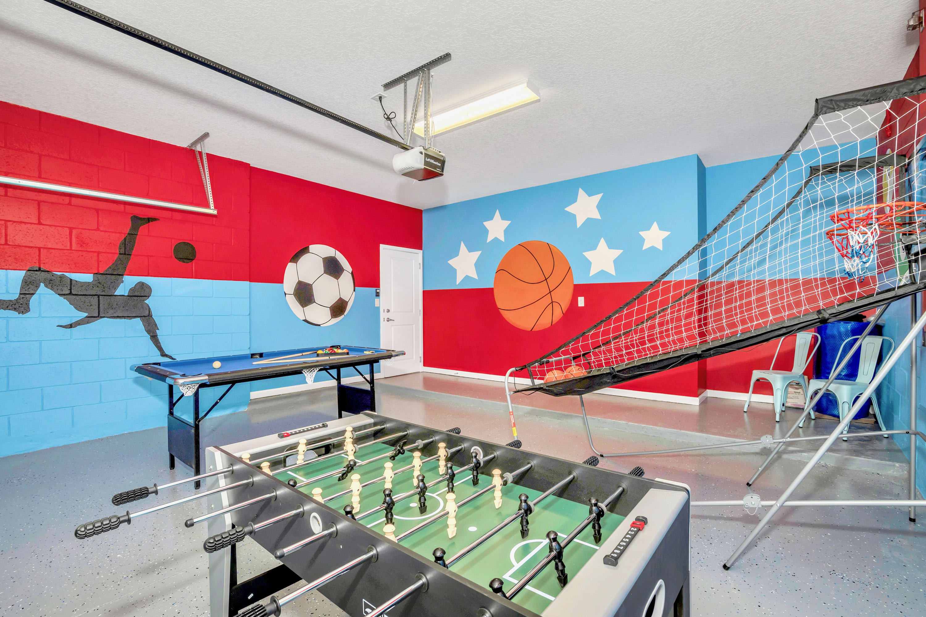 Vibrant game room in a garage with red and blue walls featuring sports-themed murals. It includes a blue felt pool table, a foosball table, and a basketball arcade game, all on a speckled gray floor.