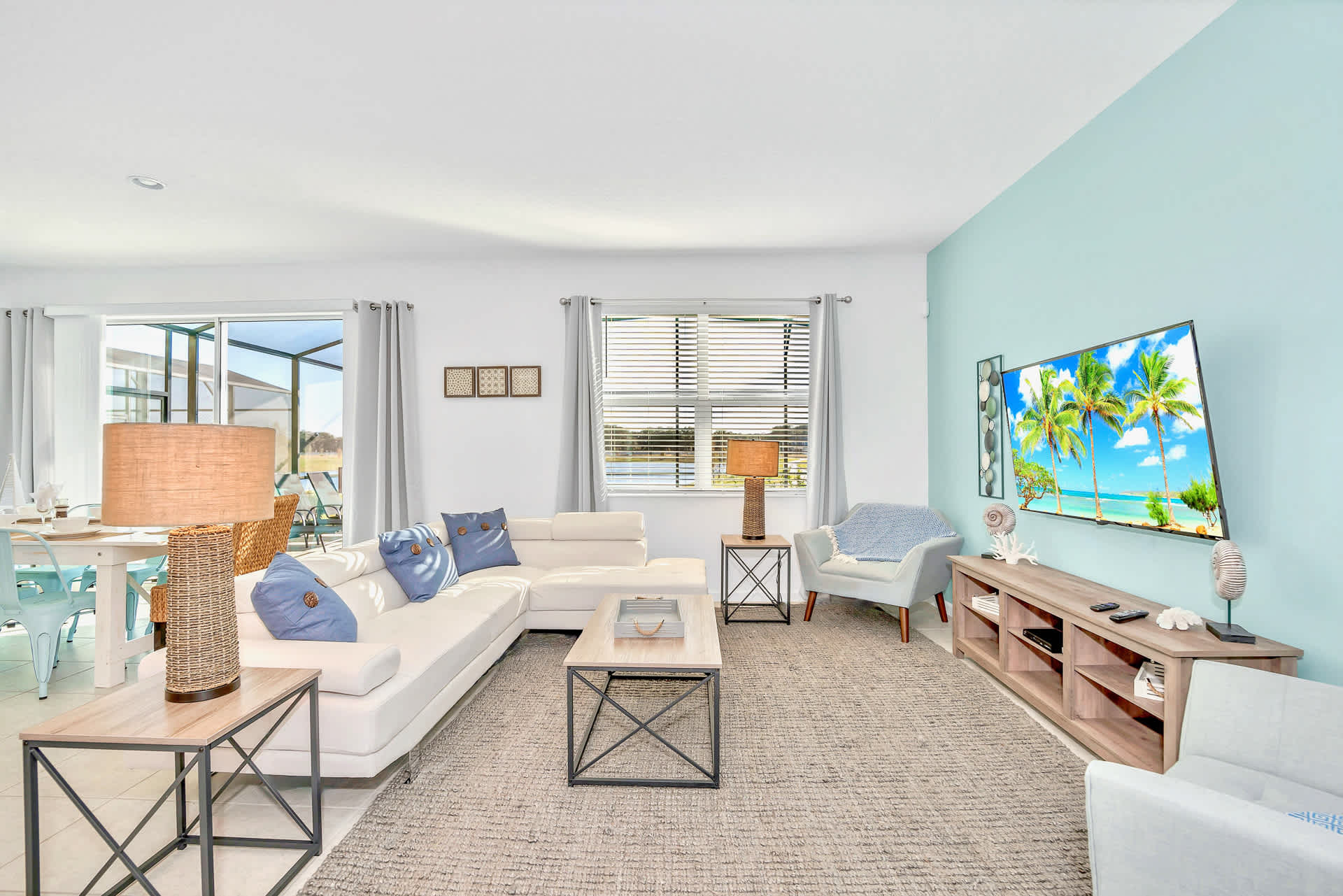 Bright living room with a white sectional sofa, a light blue accent wall, and a large flat-screen TV mounted above a wooden console. The room includes a textured rug, modern side tables, and a view of the outdoor pool area through windows.