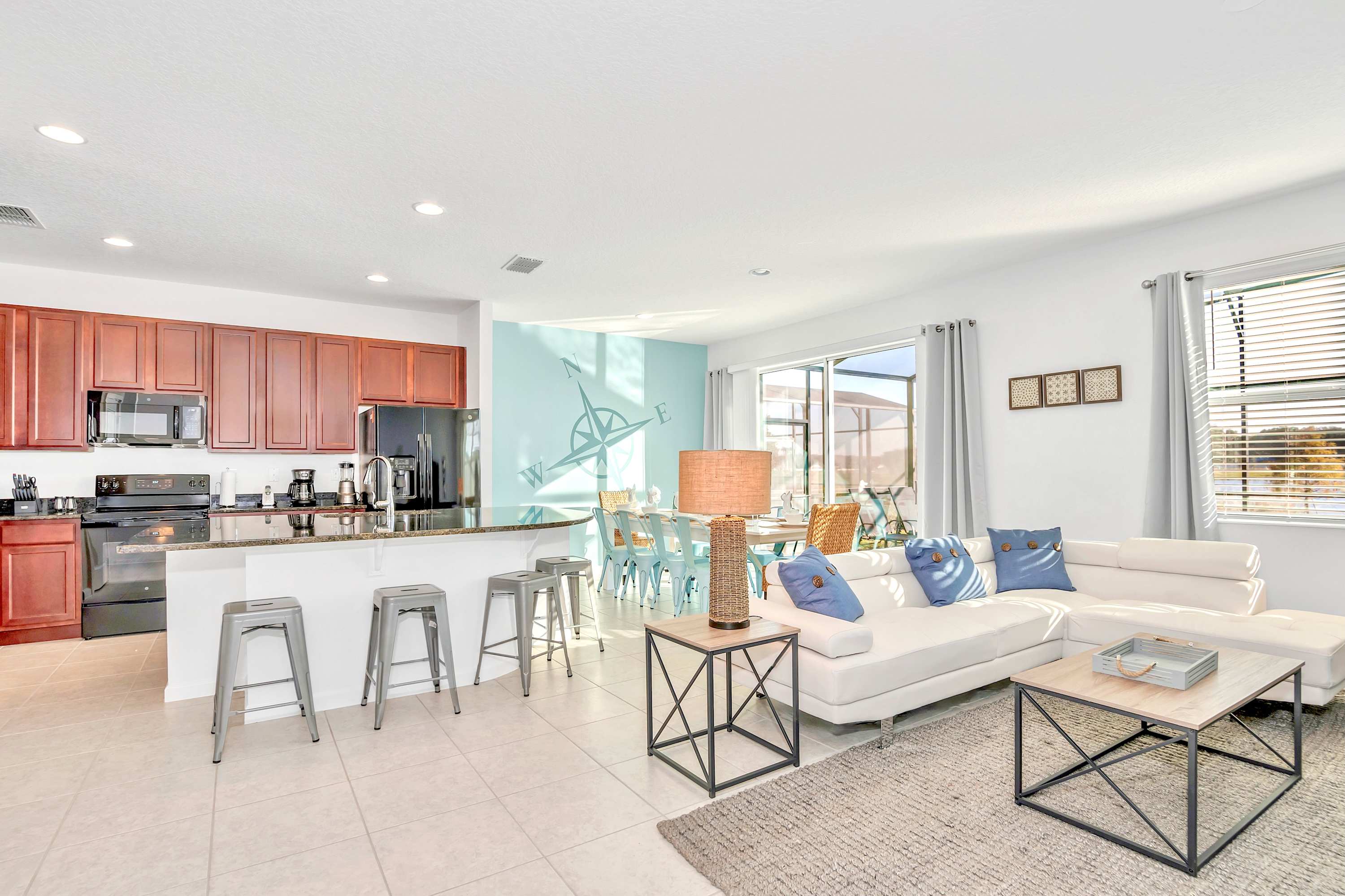 Bright open-concept living area featuring a white sectional sofa with blue pillows, modern coffee tables, and a textured rug. A kitchen island with bar stools is to the left, and a dining area with a light blue accent wall is visible in the background.