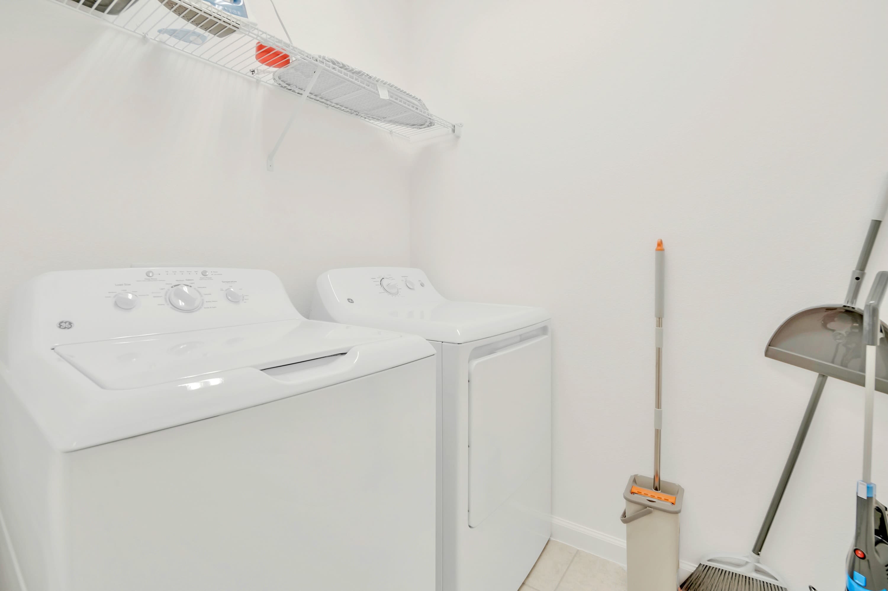 A bright white laundry room features a white top-loading washing machine and a matching dryer, with a wire shelf installed above them. Cleaning supplies like a mop and broom are neatly stored in the corner.
