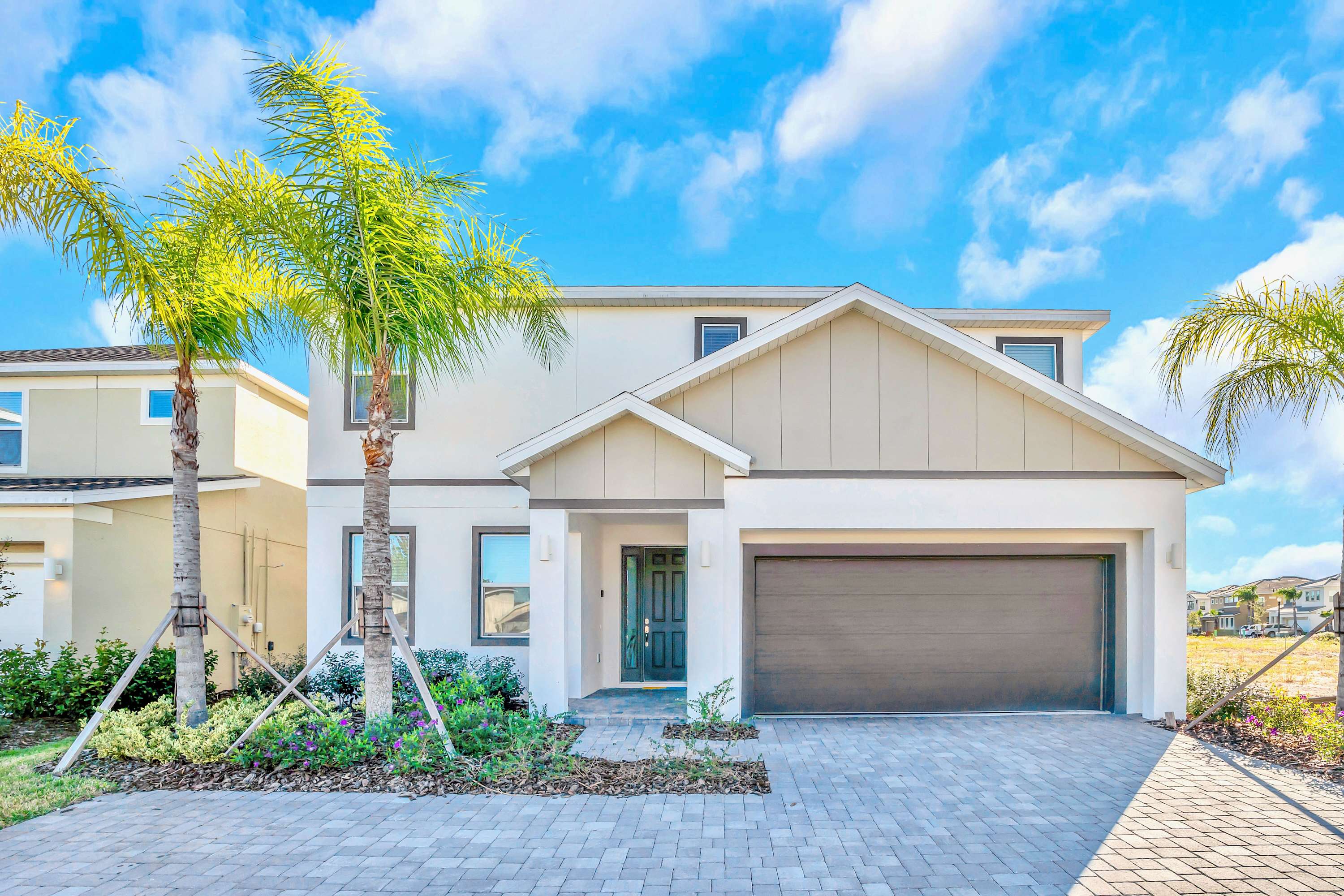 Modern two-story house exterior with a light-colored facade, dark gray garage door, and a dark front door. The front yard features a paved driveway, landscaping, and two tall palm trees under a bright blue sky.