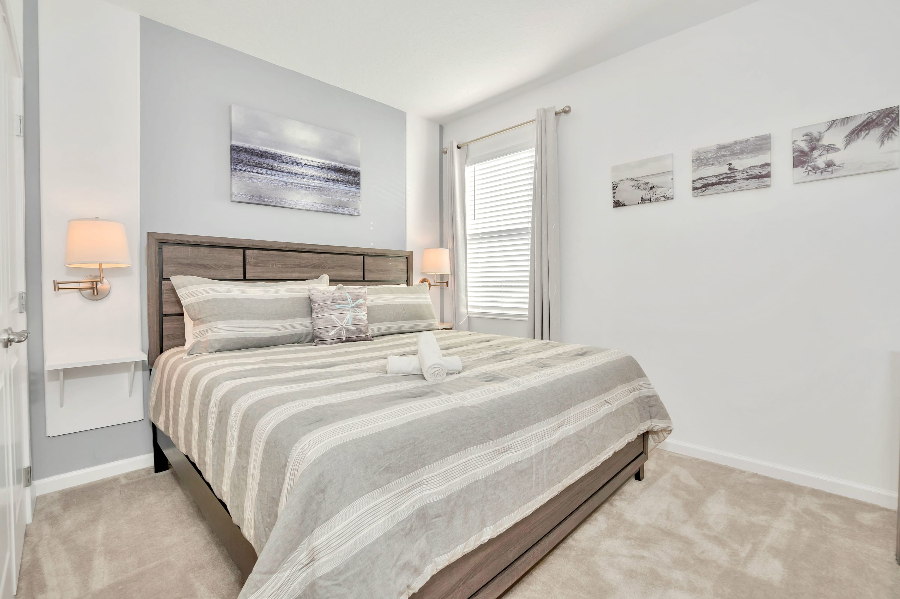 Serene bedroom featuring a large bed with grey striped bedding and a light grey accent wall, flanked by wall-mounted lights and a nightstand. A large framed seascape artwork hangs above the bed, and a window with blinds is present.