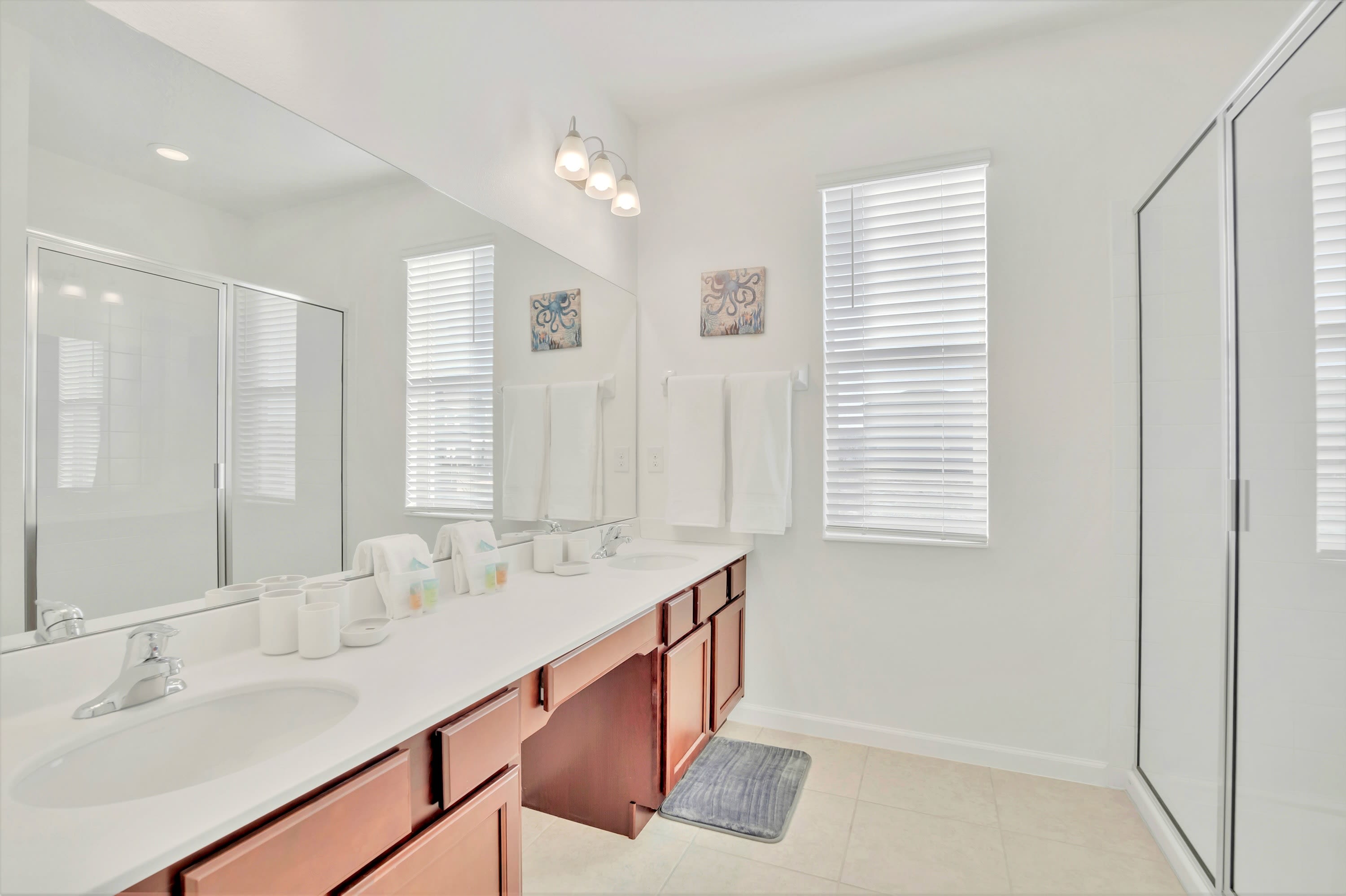 Bright bathroom featuring a long double vanity with two sinks, a large mirror, and dark wood cabinets, alongside a walk-in shower with a glass enclosure and a window with blinds.
