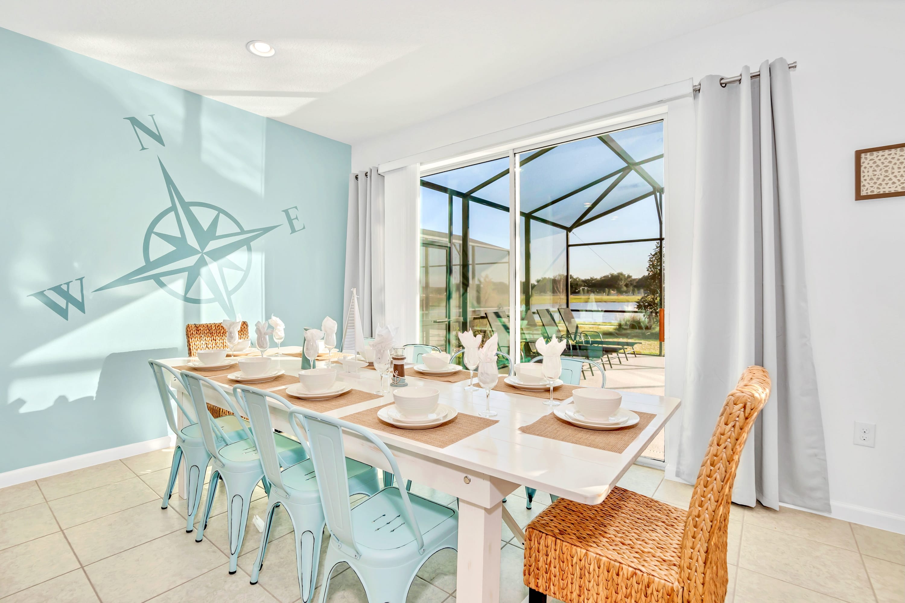 Bright dining room with a light blue accent wall featuring a large compass mural, a long white dining table with light blue chairs, and sliding glass doors leading to an outdoor pool area.