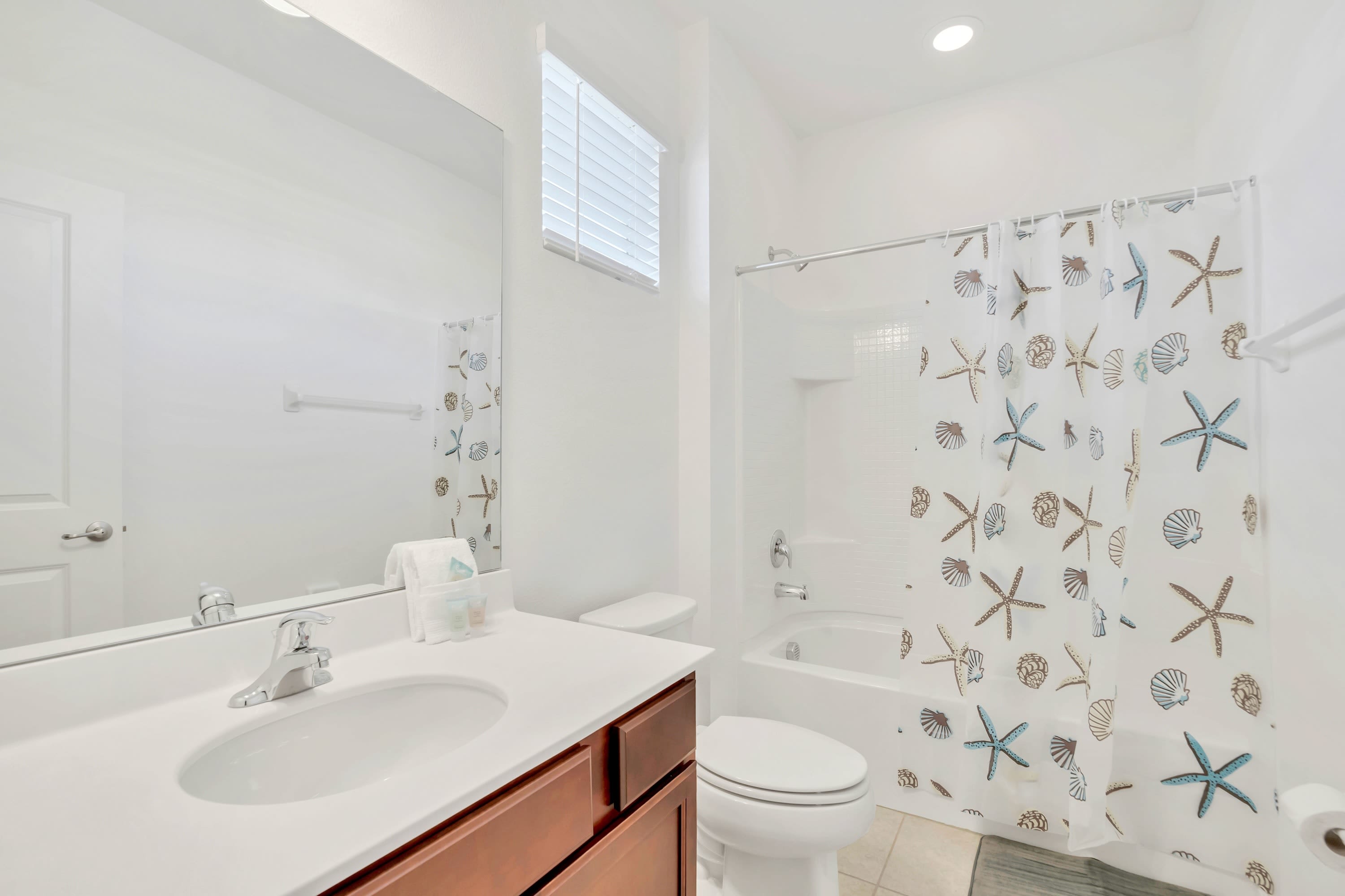 Bright bathroom with a white shower-tub combo and a starfish-patterned curtain, a toilet, and a vanity with a white countertop and wooden cabinets. A large mirror reflects the room, and a small window is above the tub.