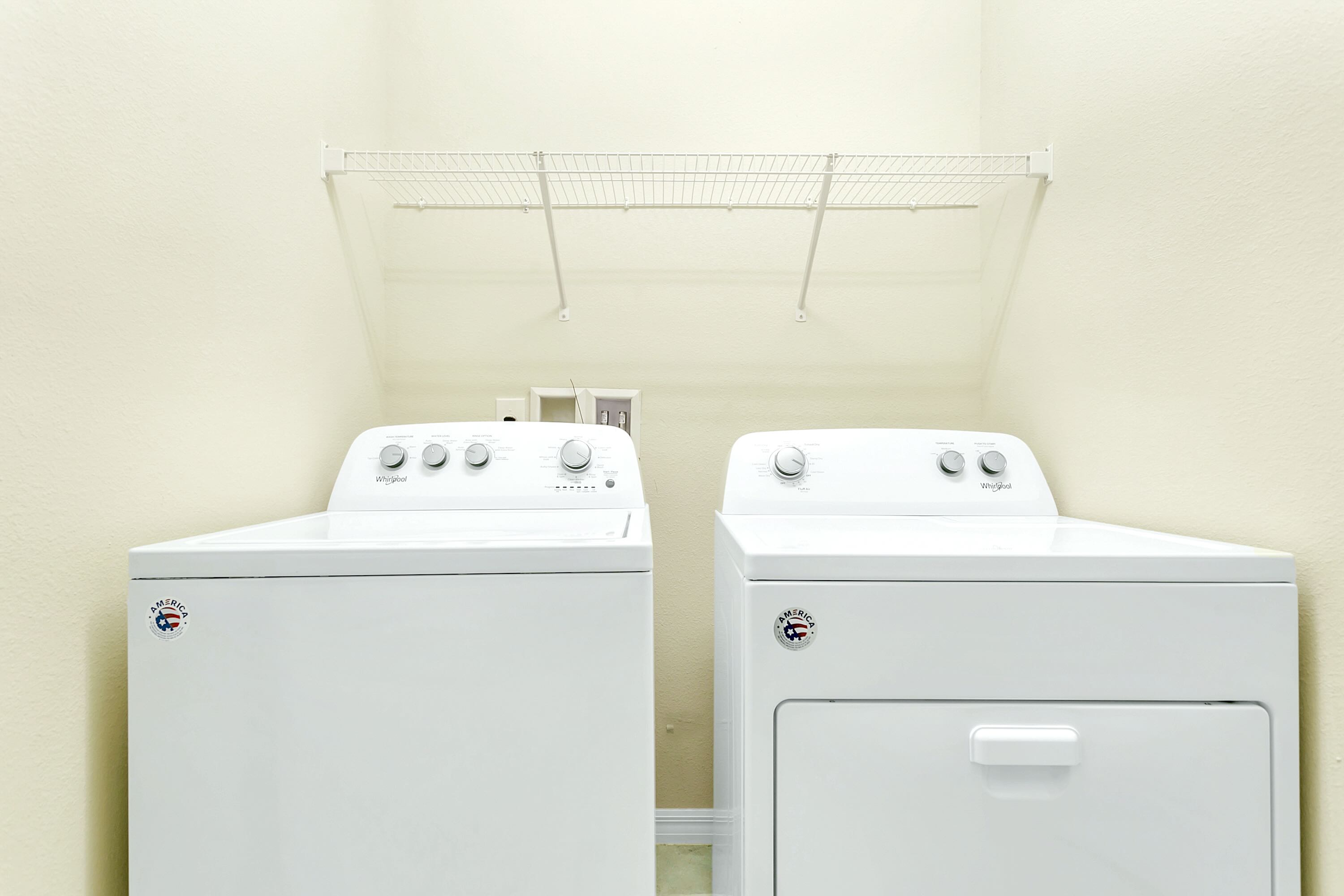 A functional laundry area equipped with a white top-loading washing machine and a front-loading dryer, positioned beneath a wire shelf against light-colored walls.