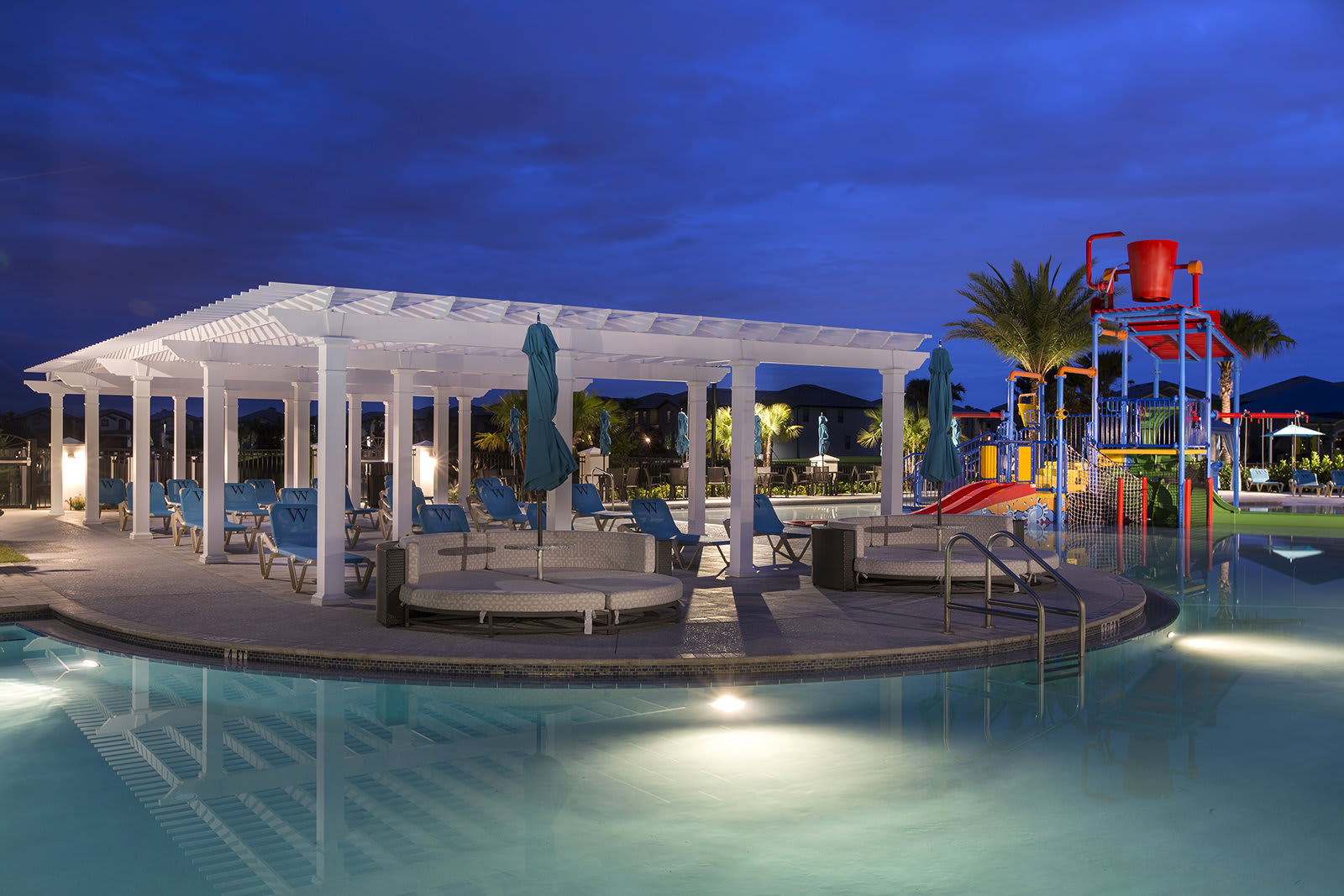 The communal pool area at dusk, illuminated by underwater lights and spotlights, showcasing a large swimming pool, a white pergola with lounge seating, and the colorful children's splash pad in the background. The sky is a deep blue with hints of twilight.