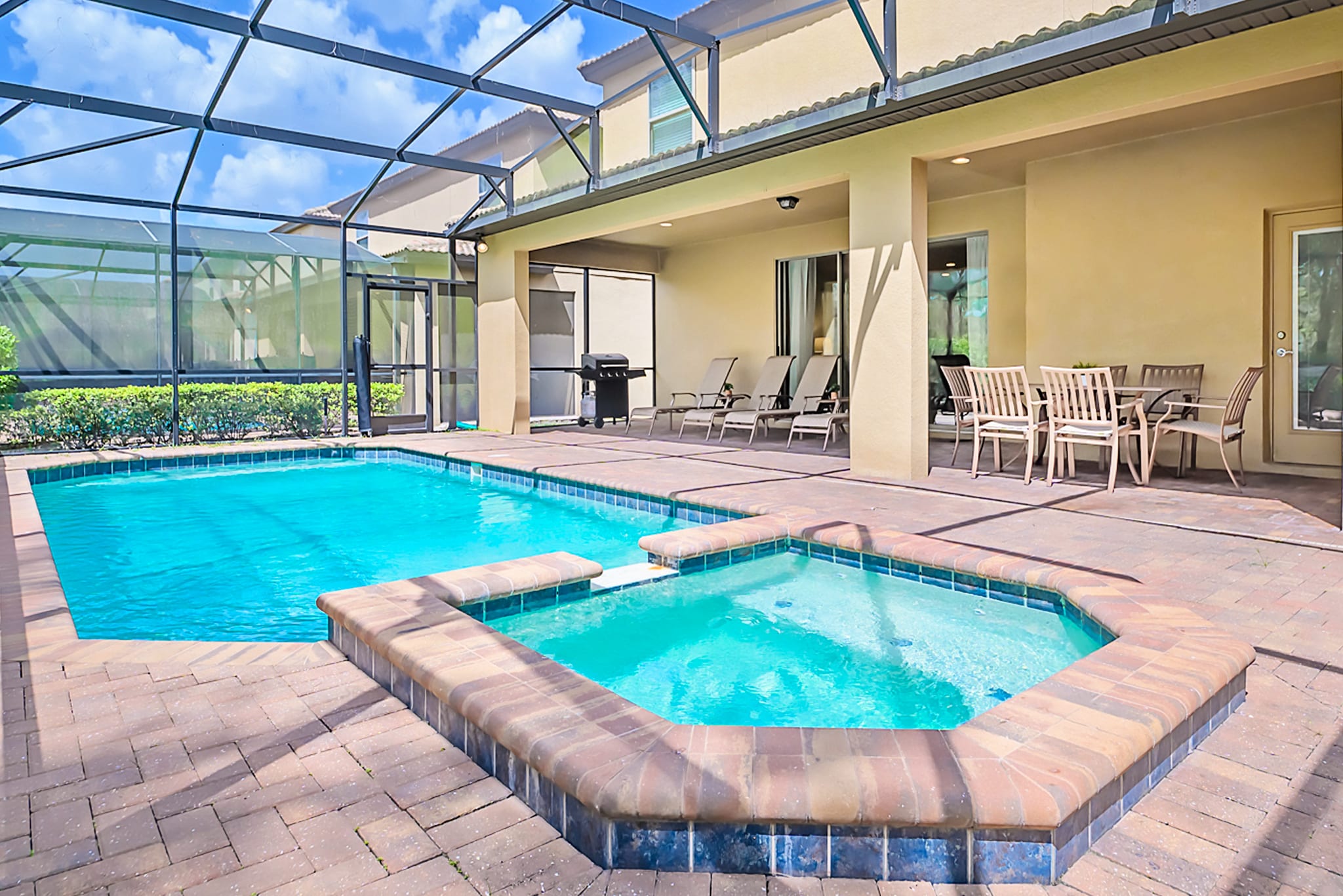 Screened-in outdoor patio featuring a rectangular private swimming pool with a connected hot tub, surrounded by brick pavers and lounge chairs, with a covered dining area and BBQ grill.