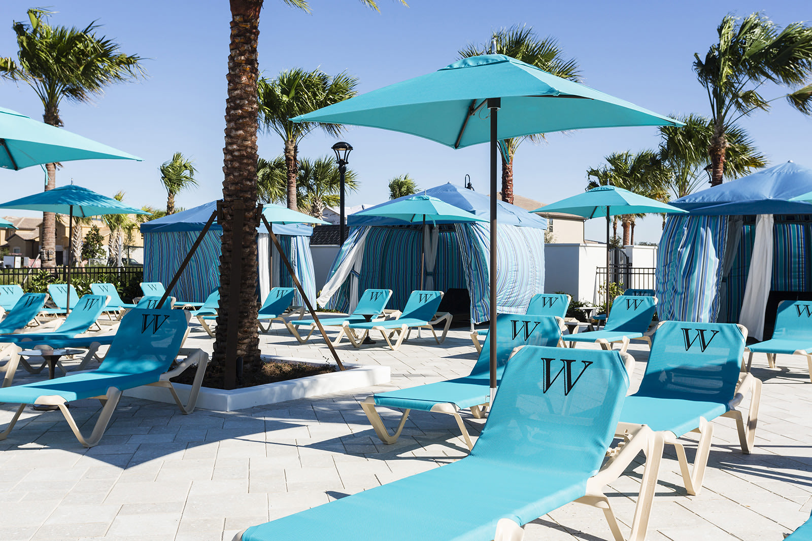A sunny pool deck area furnished with numerous turquoise lounge chairs, many featuring a 'W' logo, and several striped blue and white cabanas offering shade. Tall palm trees are scattered throughout the paved area.